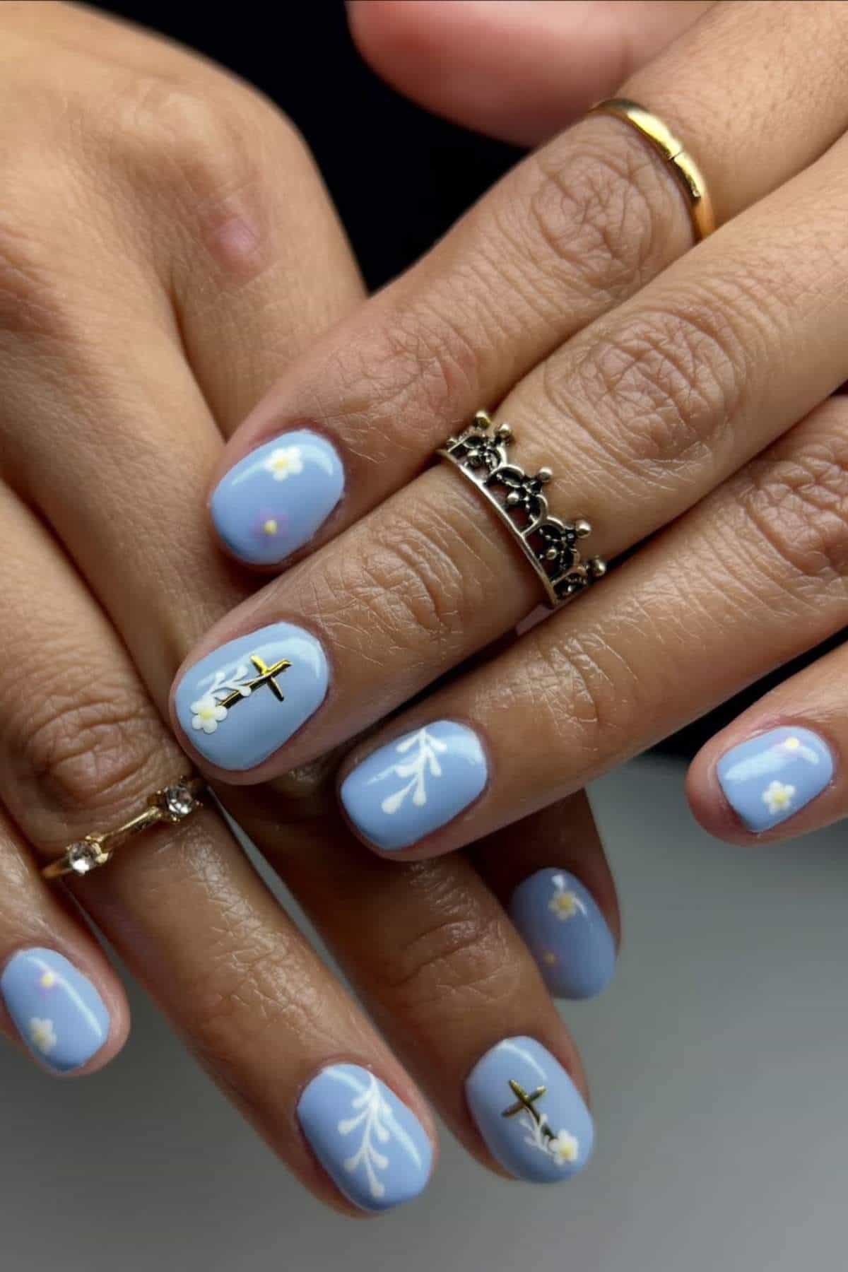 Close-up of hands with light blue nail polish, featuring small white flower designs and black cross accents on several nails. The person is wearing gold and silver rings.
