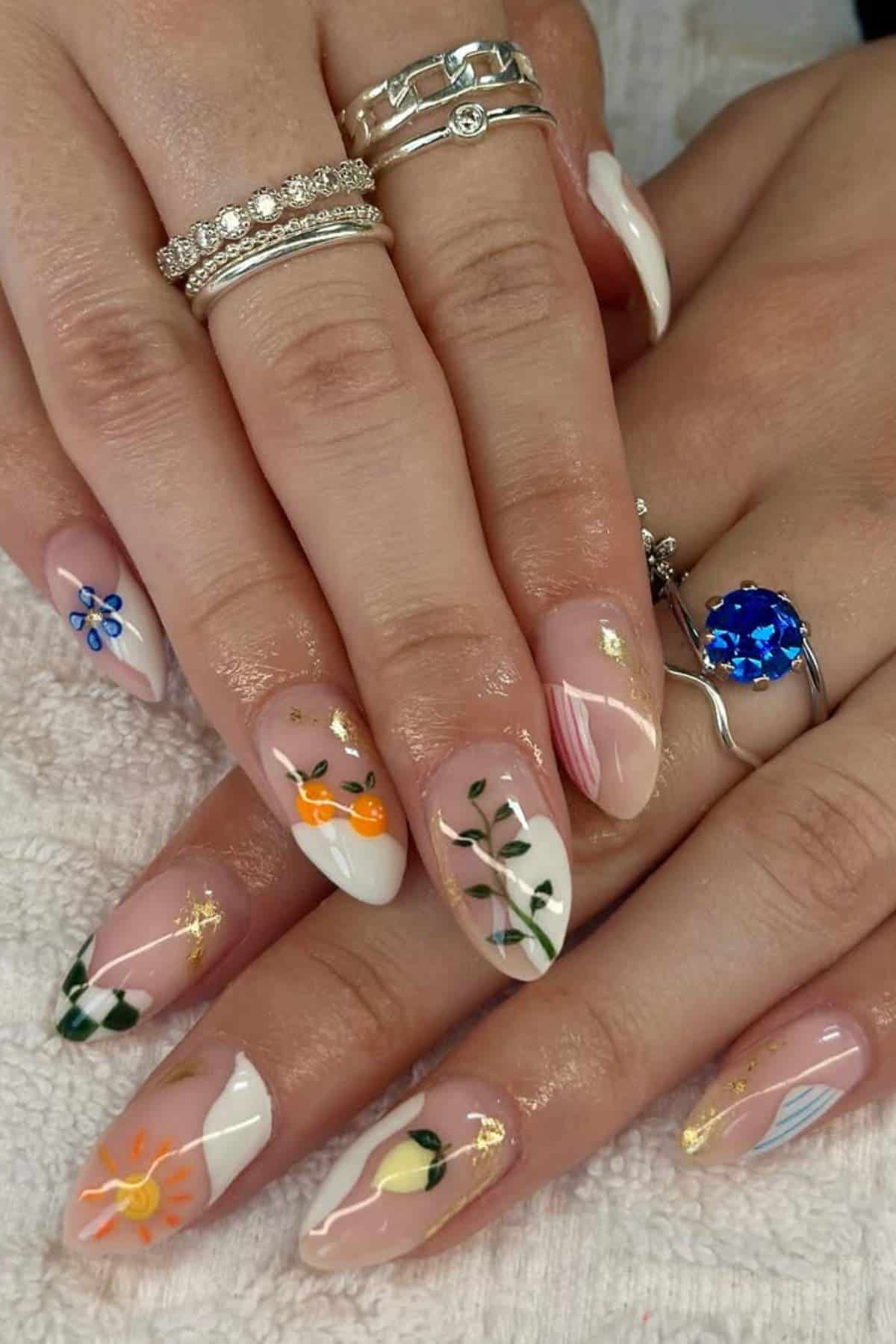 Hands with long almond-shaped nails featuring various floral and fruit designs, pastel colors, and gold accents. The person is wearing multiple silver rings.