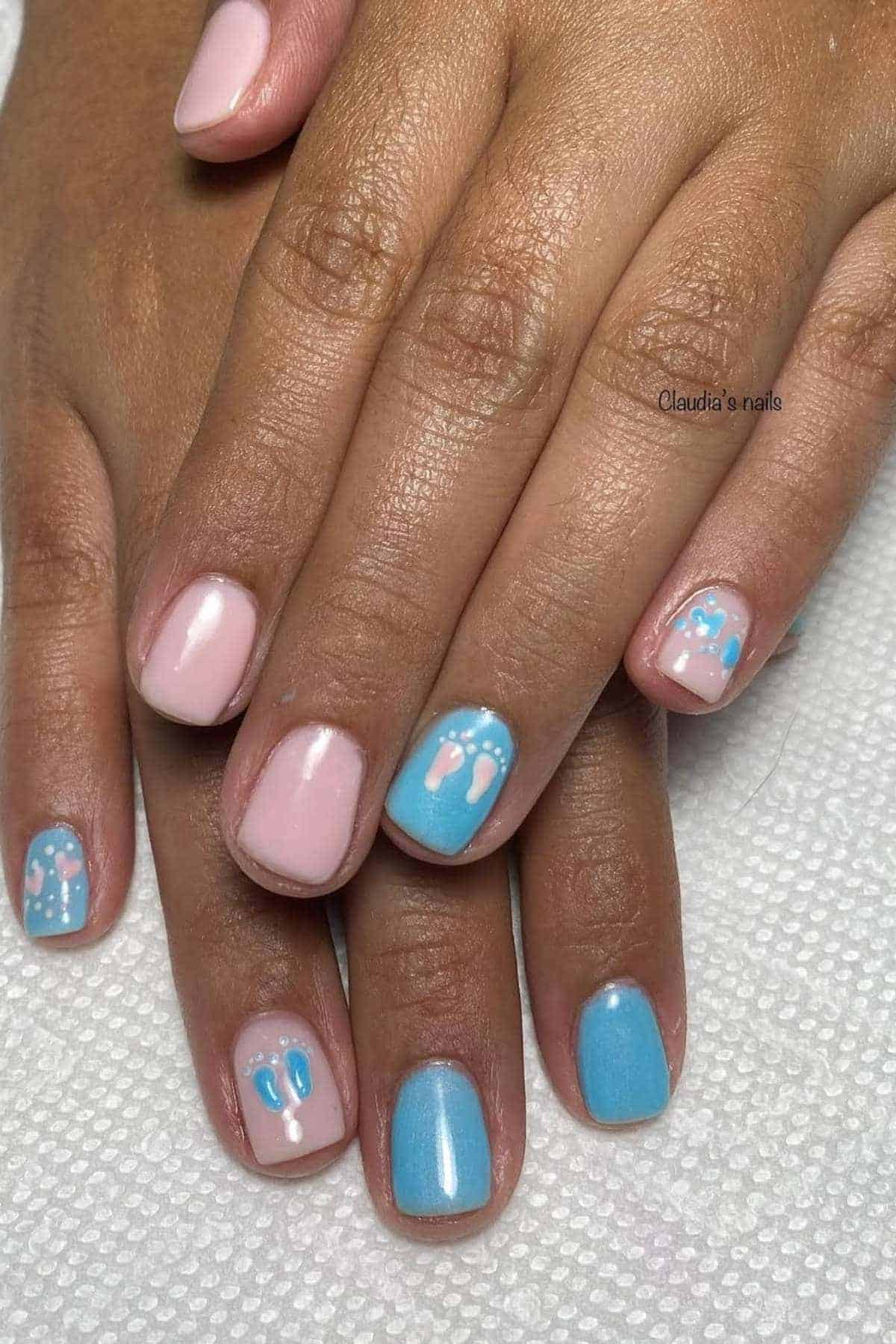Hands with short nails painted in pastel pink and blue polish, featuring white baby footprints and polka dot designs as nail art accents, resting on a white textured surface.