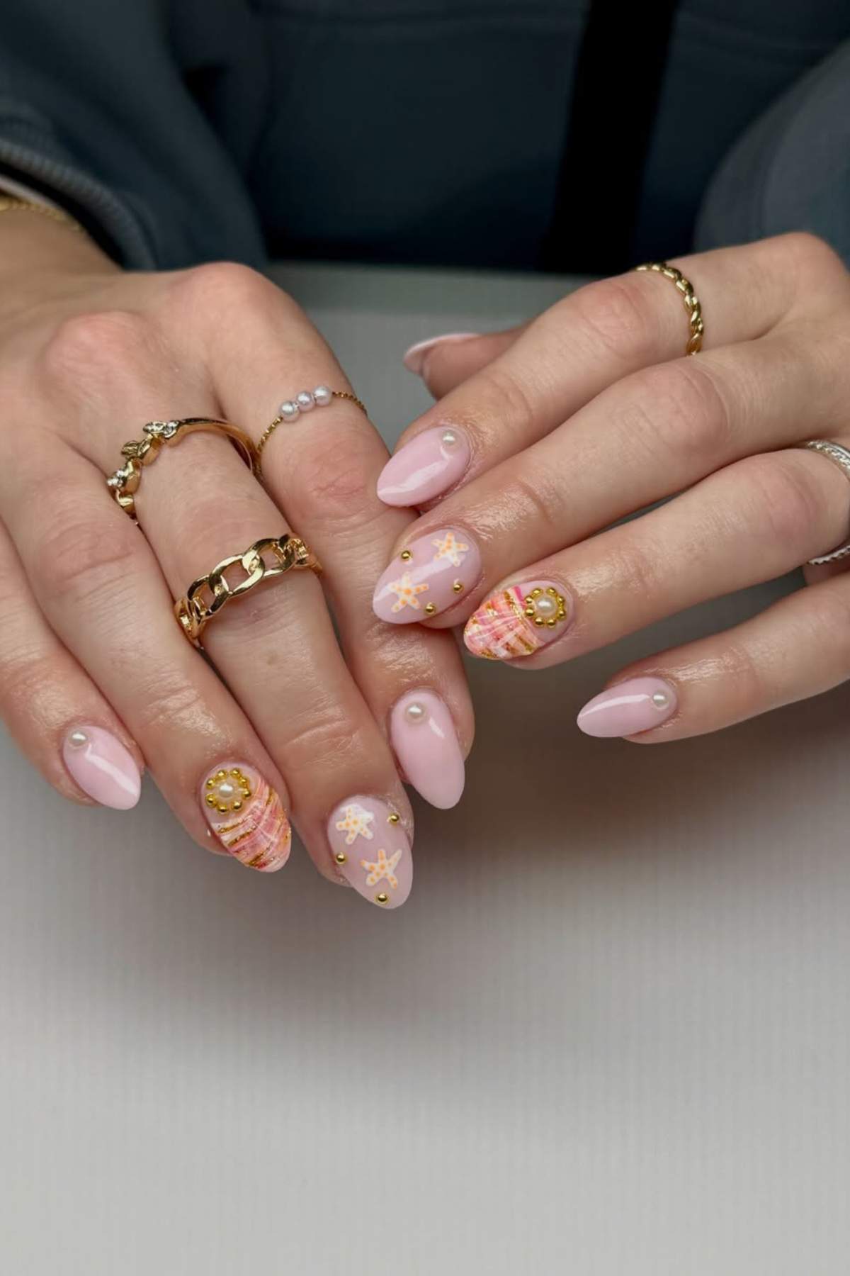 Hands with almond-shaped nails featuring pink and peach nail art, gold accents, and pearl embellishments. The person is wearing multiple gold and pearl rings.