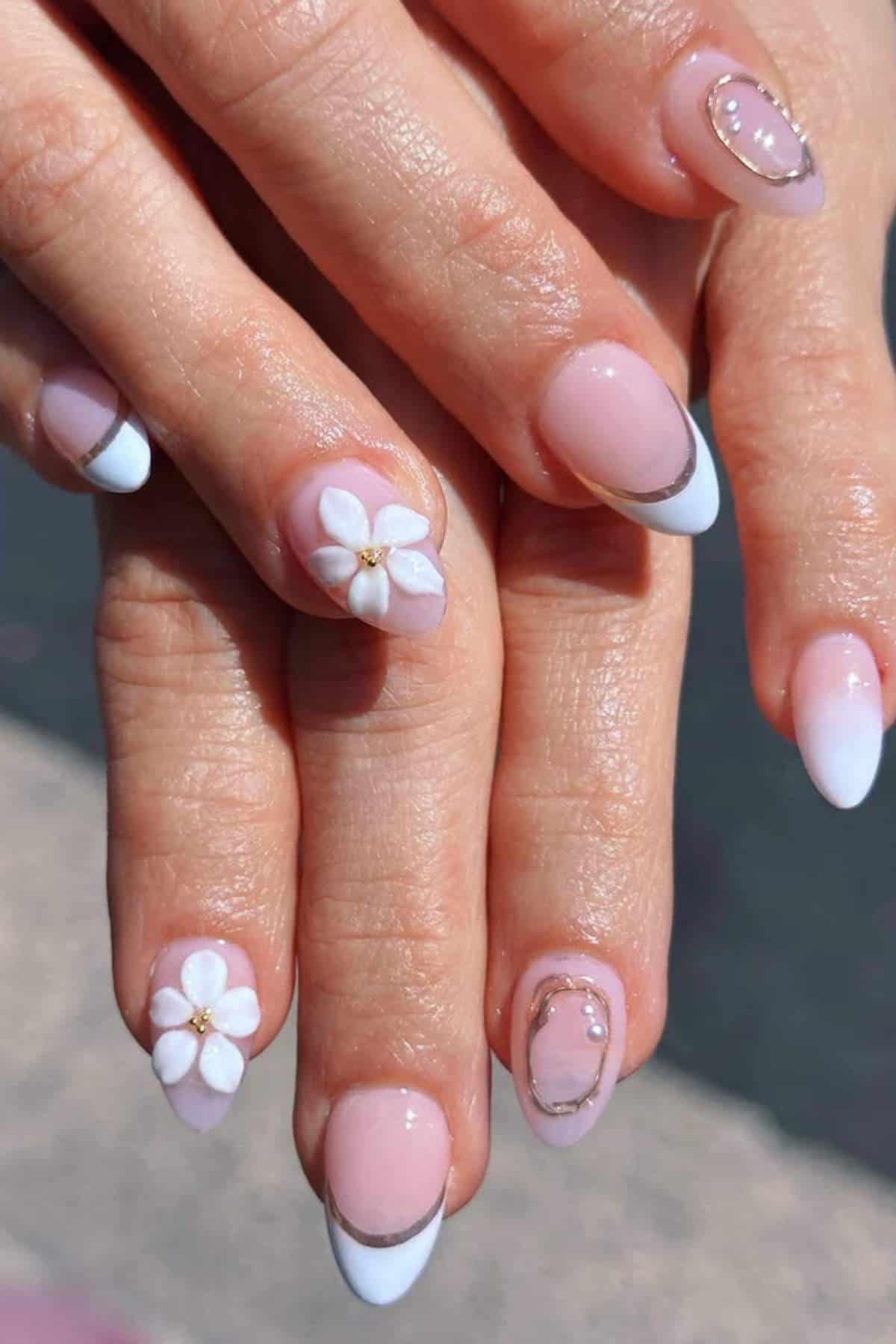 A close-up of hands with almond-shaped nails featuring a pink and white French tip design, floral 3D decorations, and gold embellishments.