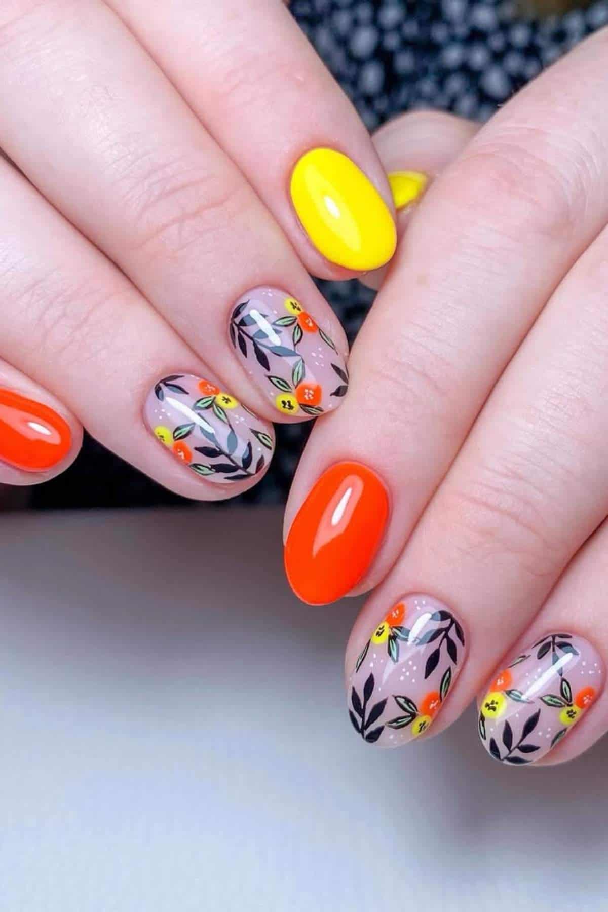 Close-up of hands with manicured nails, featuring bright yellow and orange polish, and floral designs with black leaves and small orange and yellow flowers on some nails.