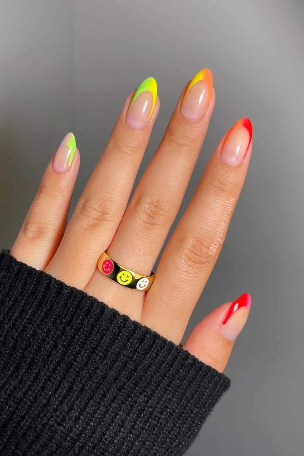 A hand with almond-shaped nails features a gradient colorful French tip manicure and wears a black ring with red, yellow, and white smiley faces.