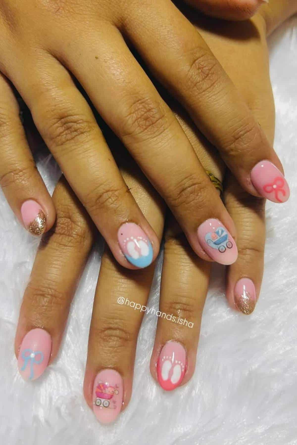 Hands with pink and gold glitter nail polish featuring designs of bows and baby feet in blue and white on a white textured surface.