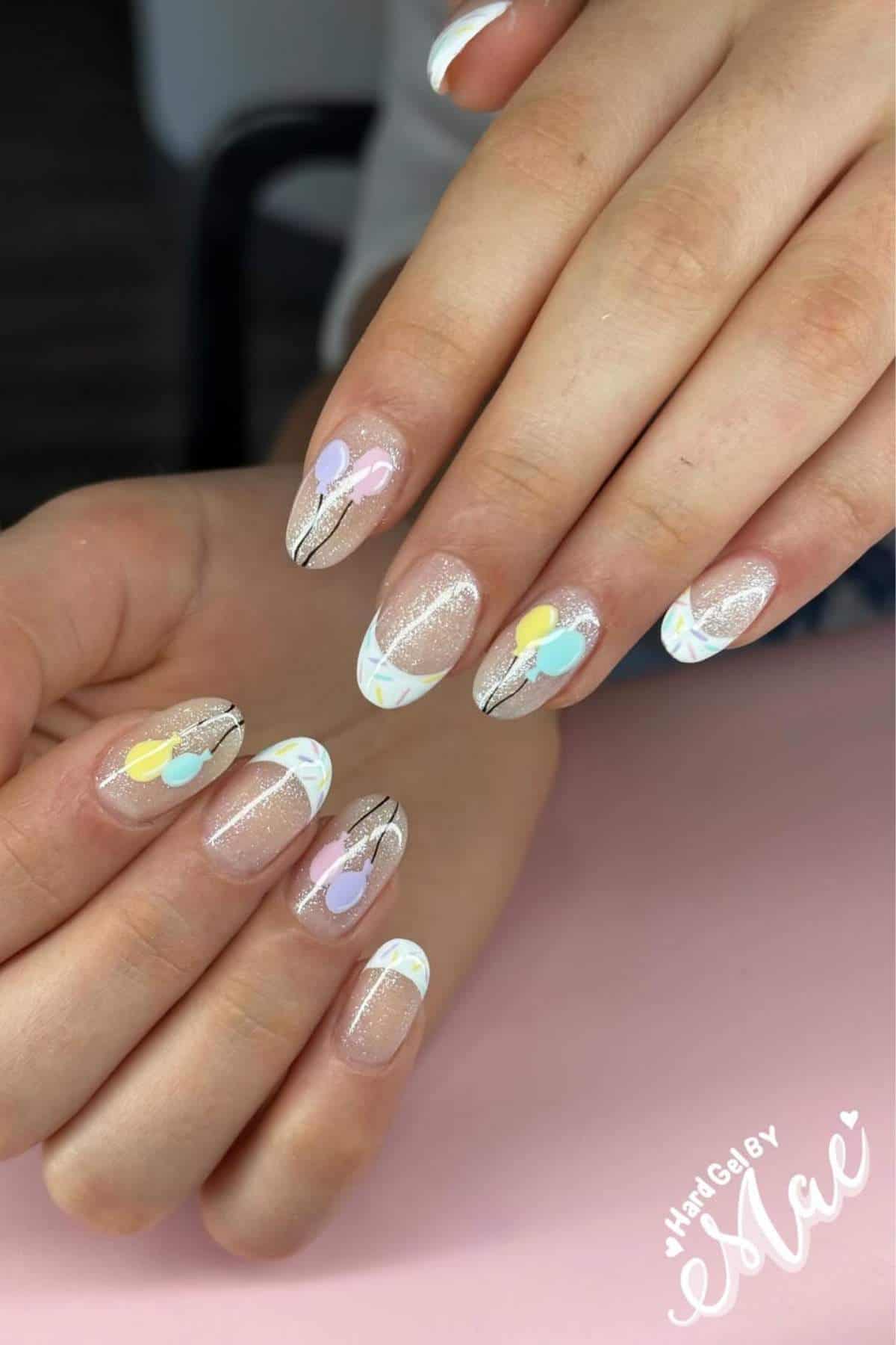 Hands with almond-shaped nails featuring pastel balloon designs, white French tips, and glitter accents on a light pink surface.