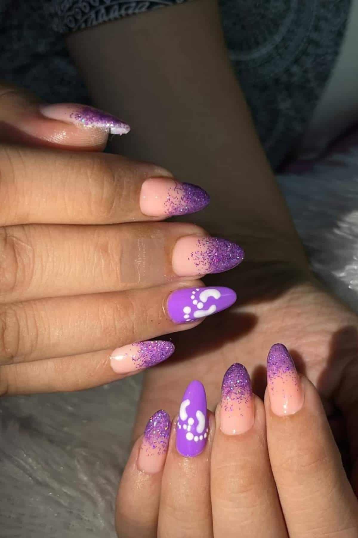 Hands with almond-shaped nails featuring a pink base, purple glitter tips, and two accent nails painted purple with white dot and swirl designs.