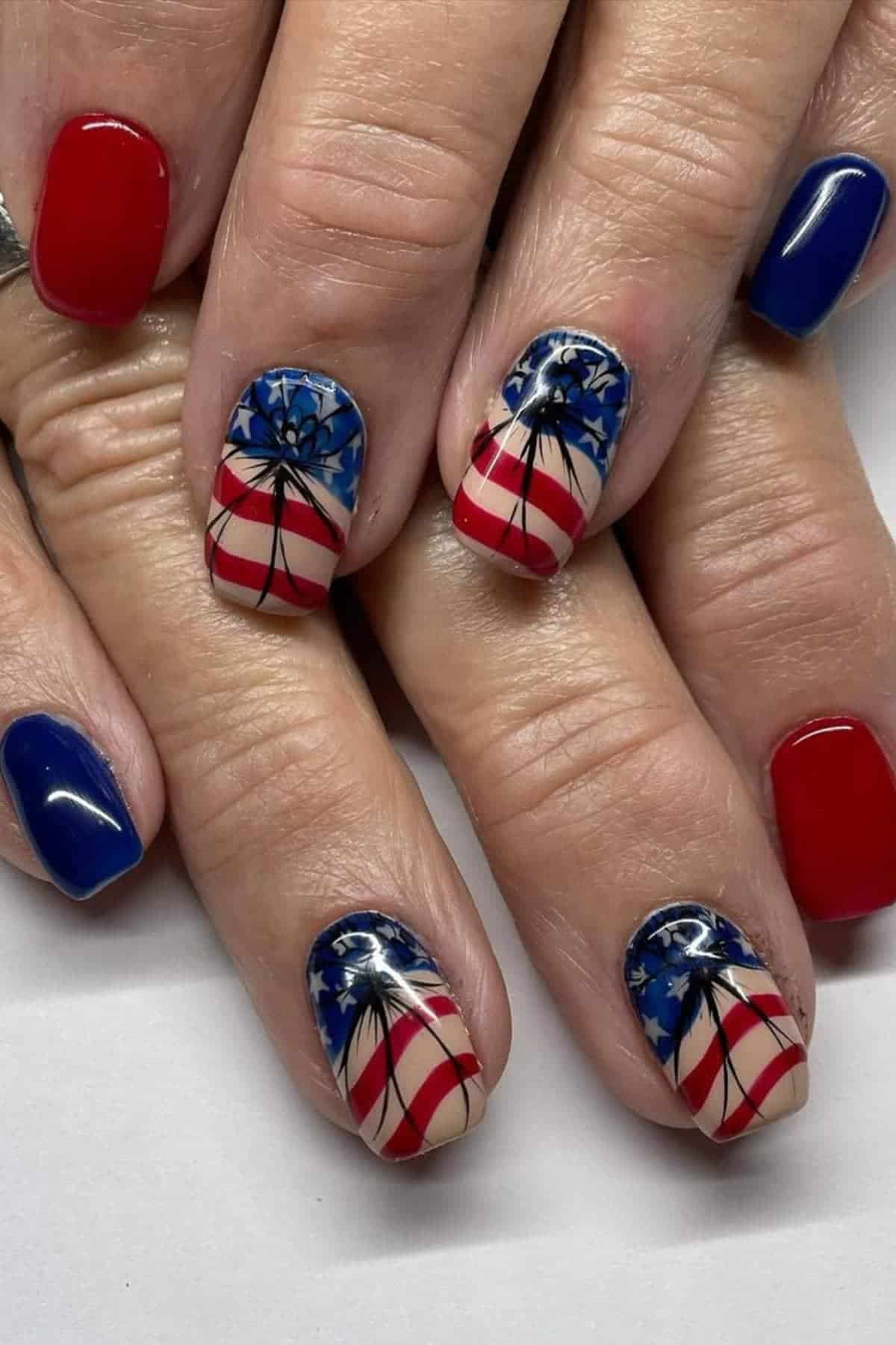 Close-up of hands with patriotic nail art featuring red, white, and blue designs, stars, stripes, and black fireworks on some nails.