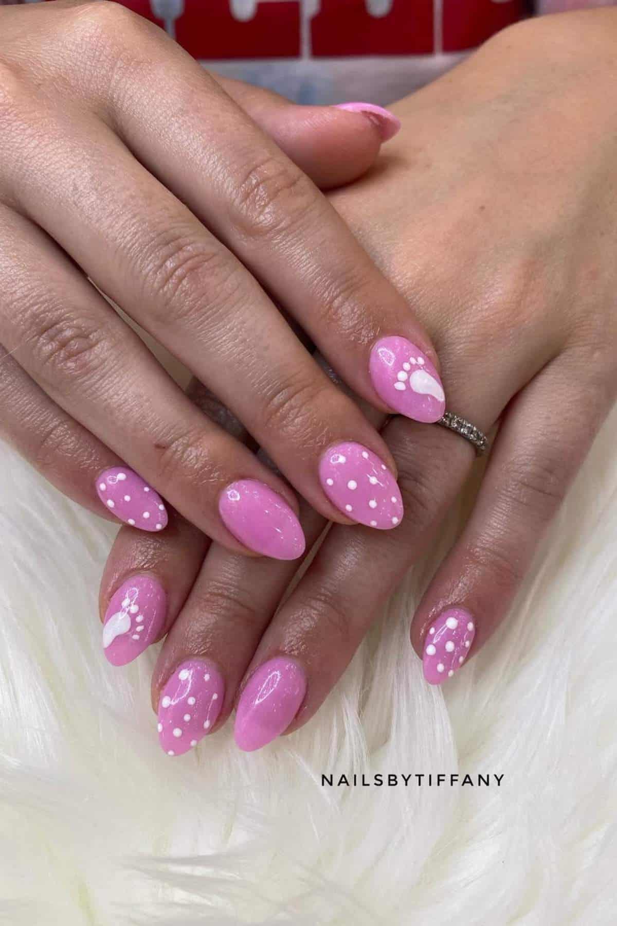 Hands with almond-shaped pink nails featuring white polka dot and heart designs, resting on a white surface.