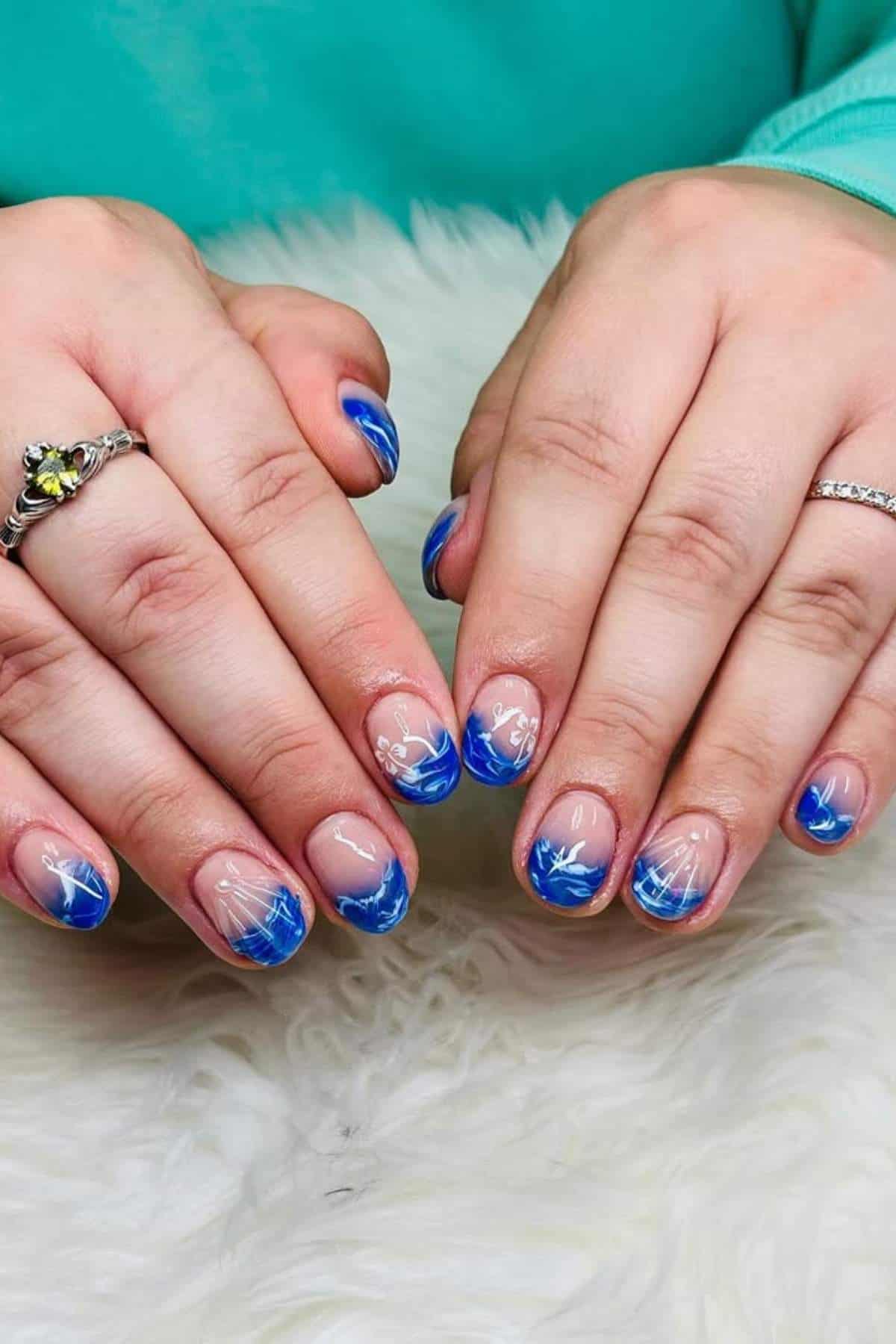 Hands with short nails featuring blue and white abstract wave designs, positioned on a white, fluffy surface. The person wears two rings and a teal top.
