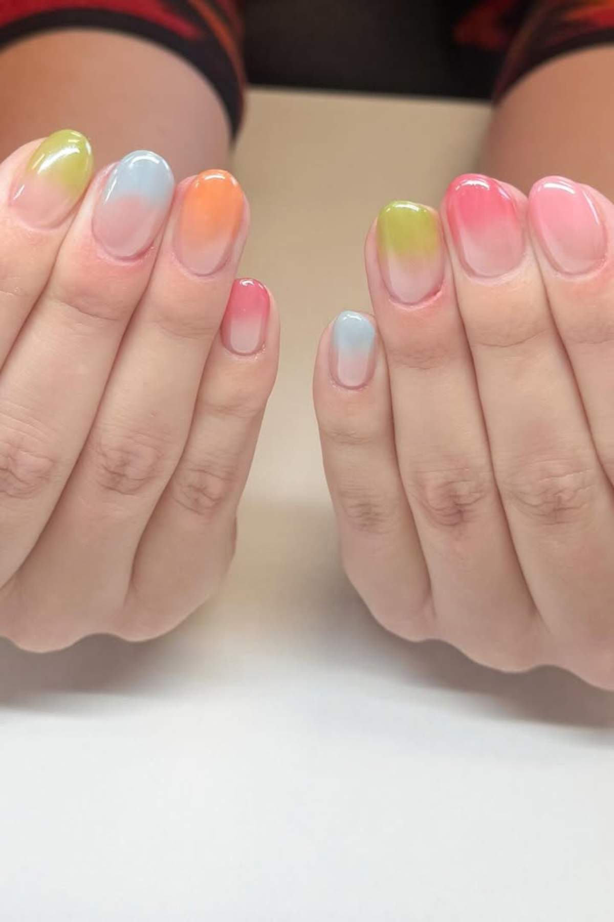 Close-up of two hands with oval-shaped nails painted in a gradient of pastel colors including pink, blue, green, and orange.