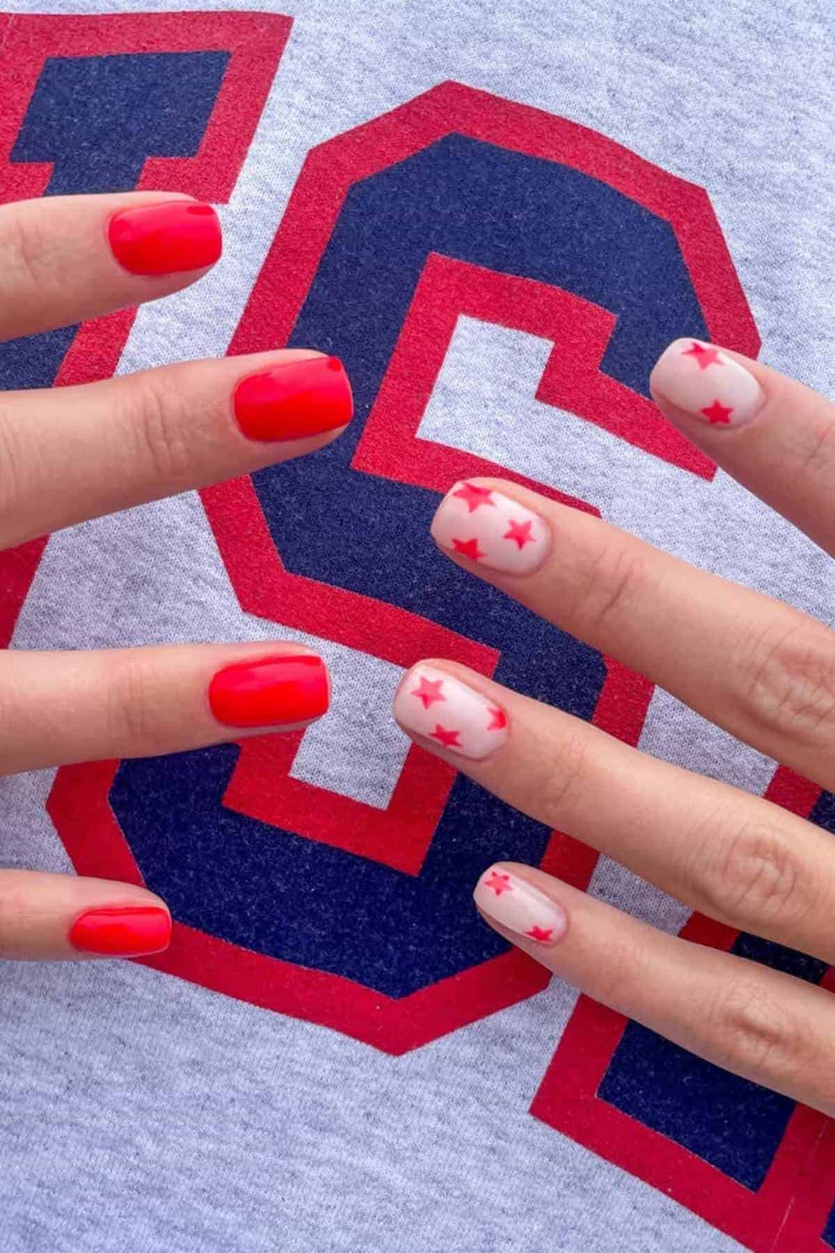 Hands with red-painted nails; two nails on each hand are painted nude with small red stars. The background is a shirt with a large red and blue "S" on it.