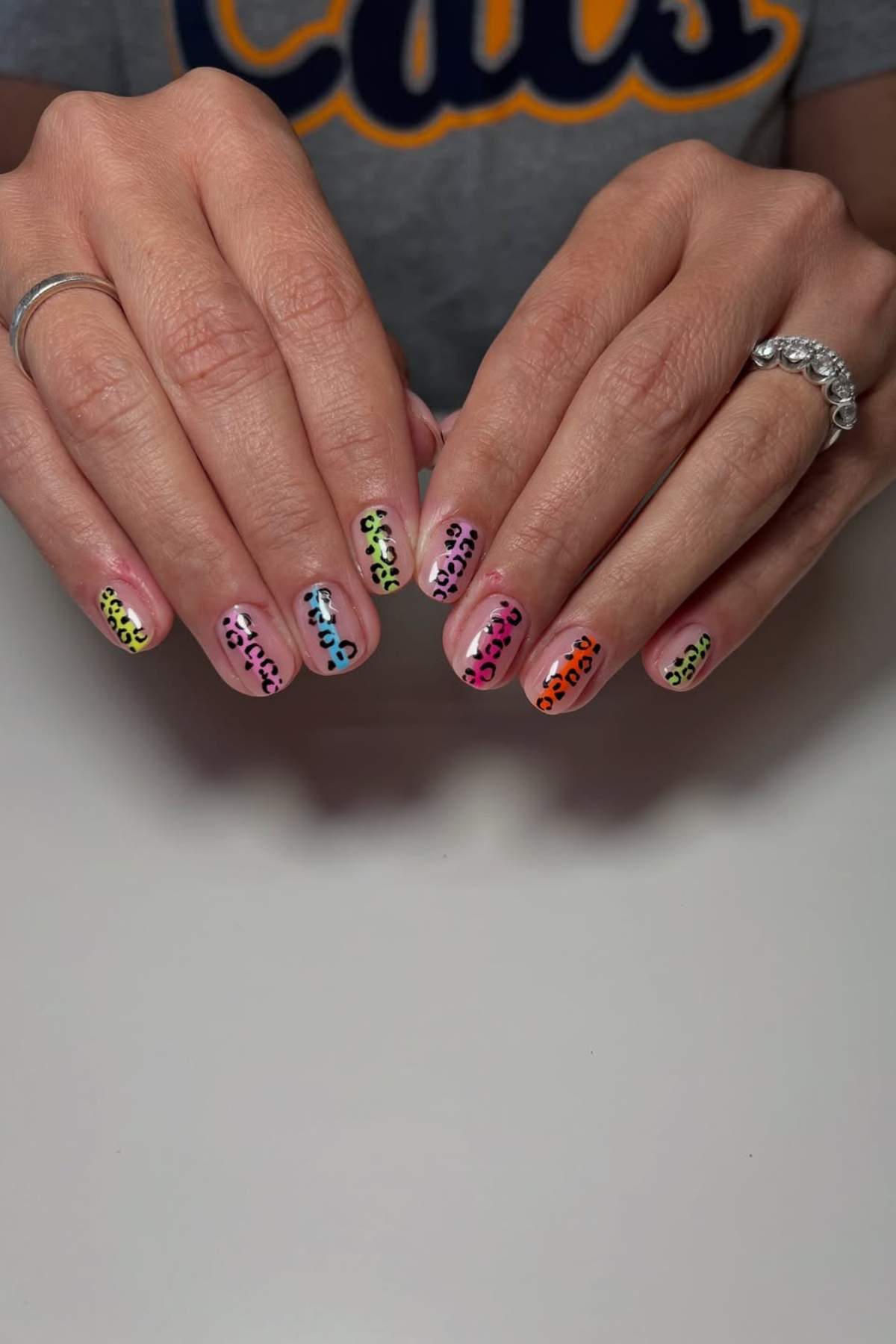 Hands with multicolored leopard print nail art on short nails, resting on a white surface. The person is wearing rings and a grey shirt with "Cats" partially visible.