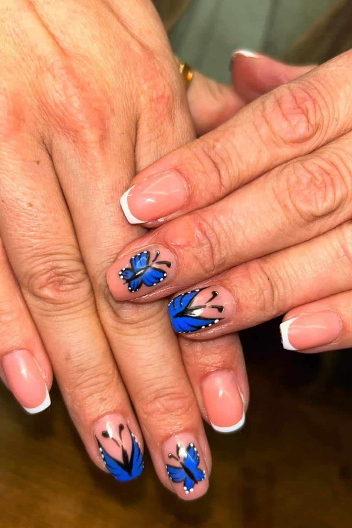 Hands with a French manicure; two nails feature blue butterfly and leaf designs on a natural nude base.