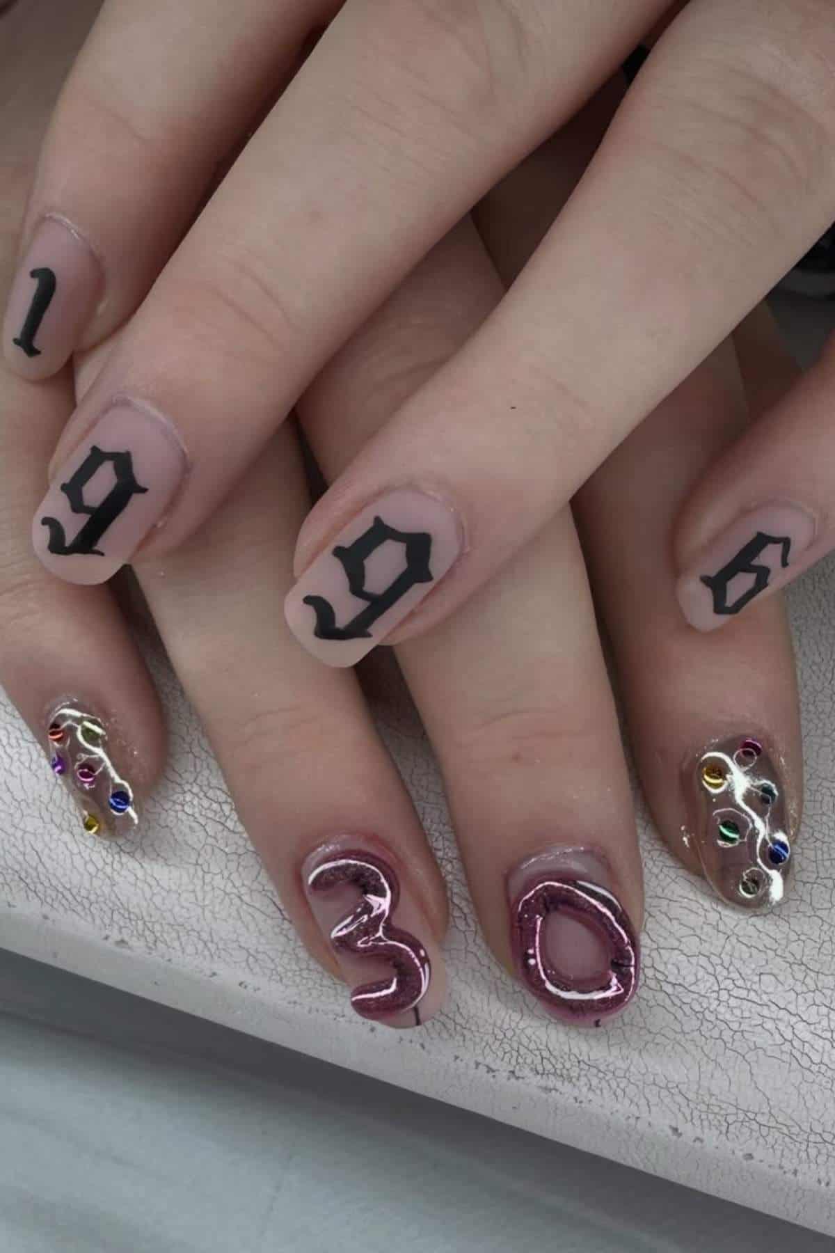 Close-up of hands with nail art; four nails display the numbers "1996" in black, while the others have colorful gems and the numbers "3" and "0" in raised designs.