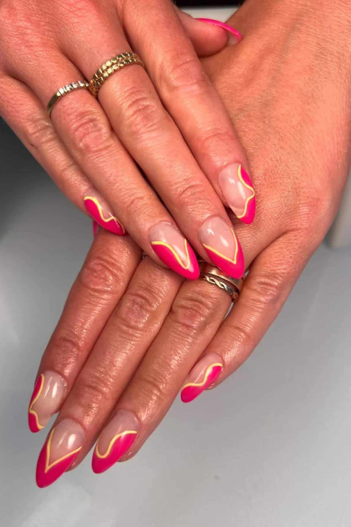 Hands with almond-shaped nails featuring a pink and yellow abstract design, with gold rings on two fingers.