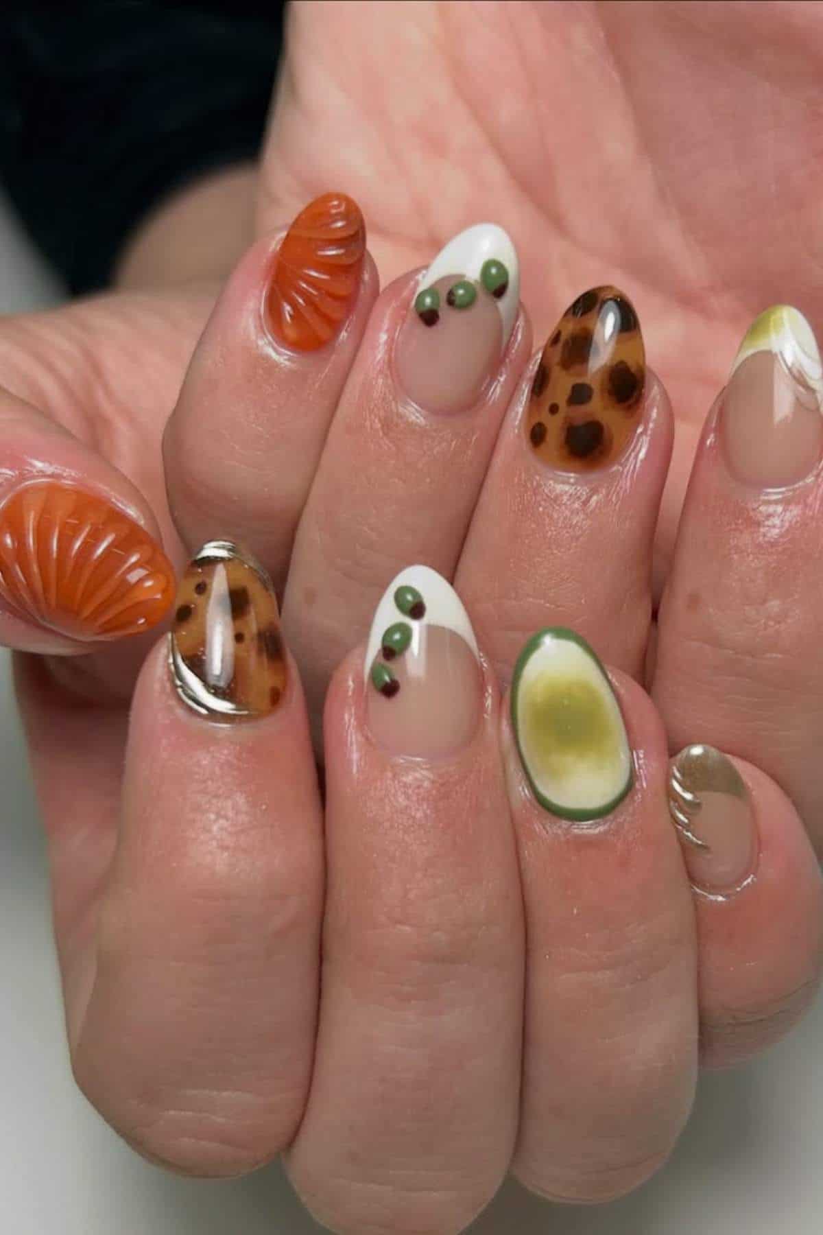 A hand with detailed nail art featuring designs resembling tortoiseshell, avocado, peas, and orange shell patterns on almond-shaped nails.