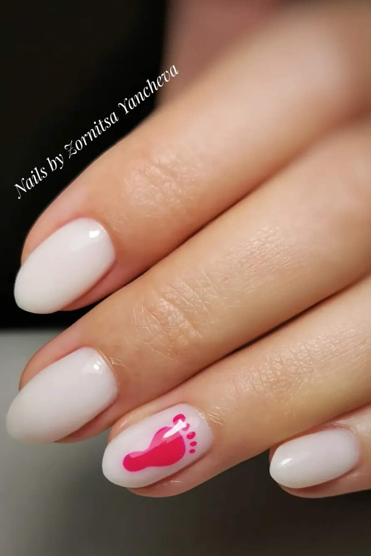 A hand with short, rounded, pale pink nails; the ring fingernail features a small pink footprint design.