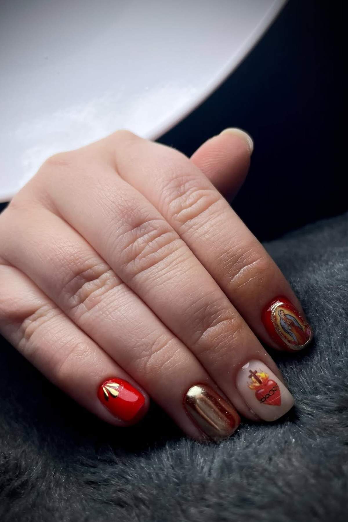 A hand with red, gold, and white nail art, including a Sacred Heart design and a religious figure on the ring and pinky fingernails, resting on a dark surface.