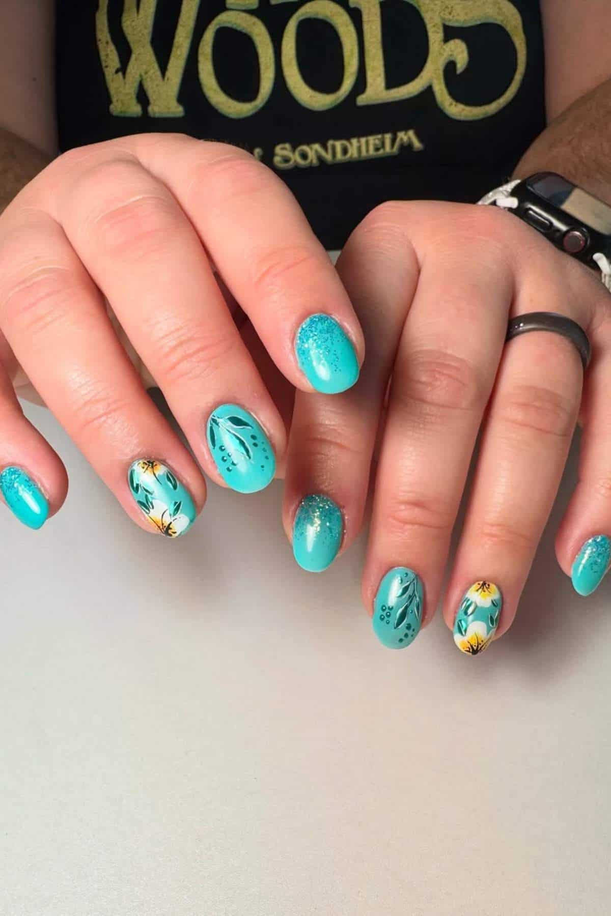 Hands with short, rounded turquoise nails featuring black, yellow, and white floral designs and some subtle glitter accents. Person wears a black shirt and a dark ring.