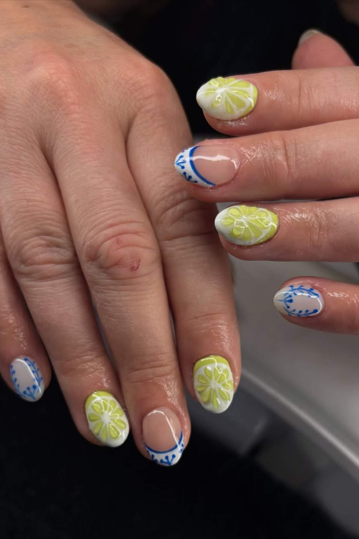 Close-up of hands with manicured nails featuring white, yellow, and blue floral and geometric designs on a short oval shape.