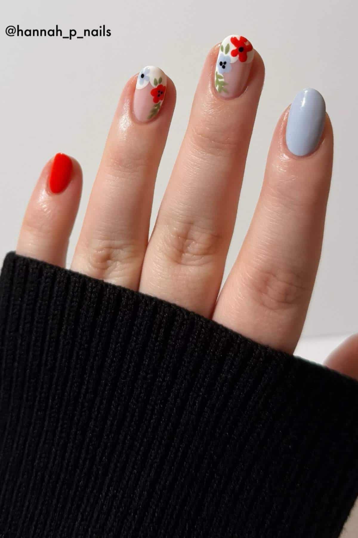 A hand with five fingernails painted in different styles, including solid red, solid light blue, and white with small floral designs. The person is wearing a black sweater.