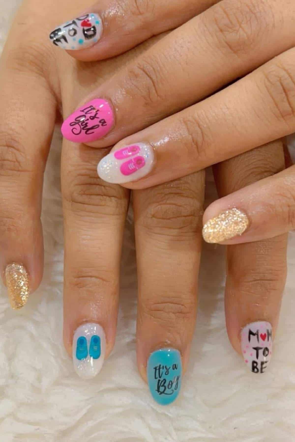 Close-up of hands with manicured nails featuring pink, blue, white, and gold polish, baby-themed designs, and phrases like "It's a Boy," "It's a Girl," and "Mom to be.