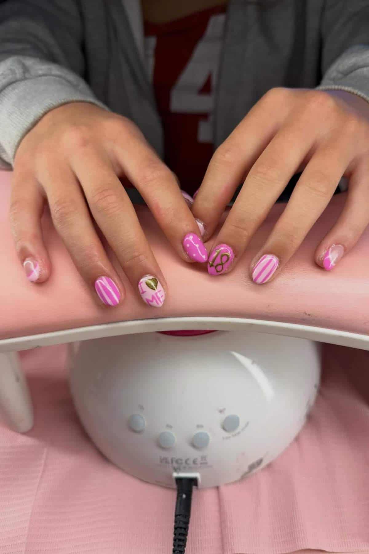 A person with manicured nails featuring pink and white designs, including stripes, hearts, and the number 18, rests their hands on a nail dryer.