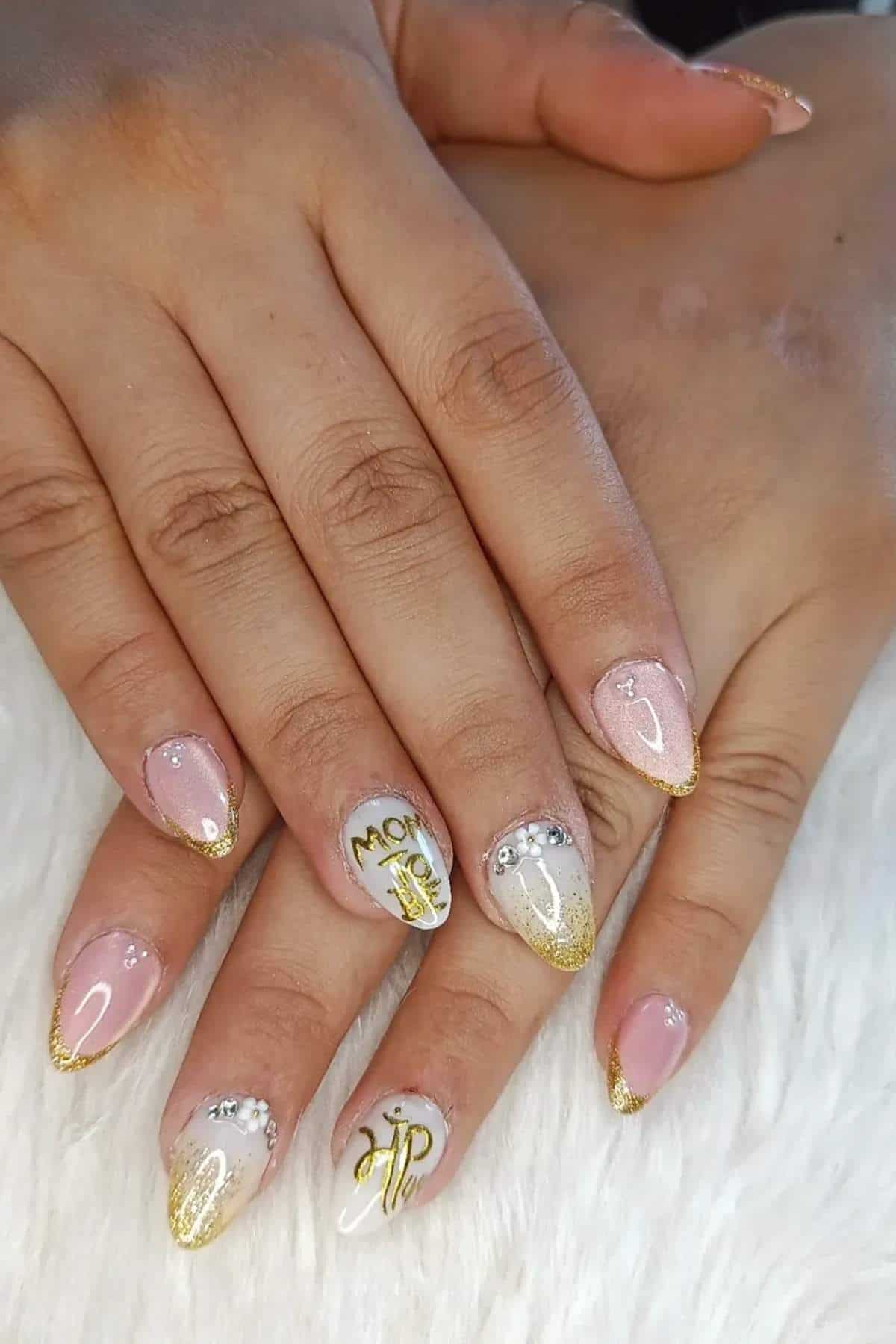 Close-up of hands with manicured nails featuring pink and white polish, gold tips, rhinestone accents, and "Mom" and "M" lettering designs on two nails.