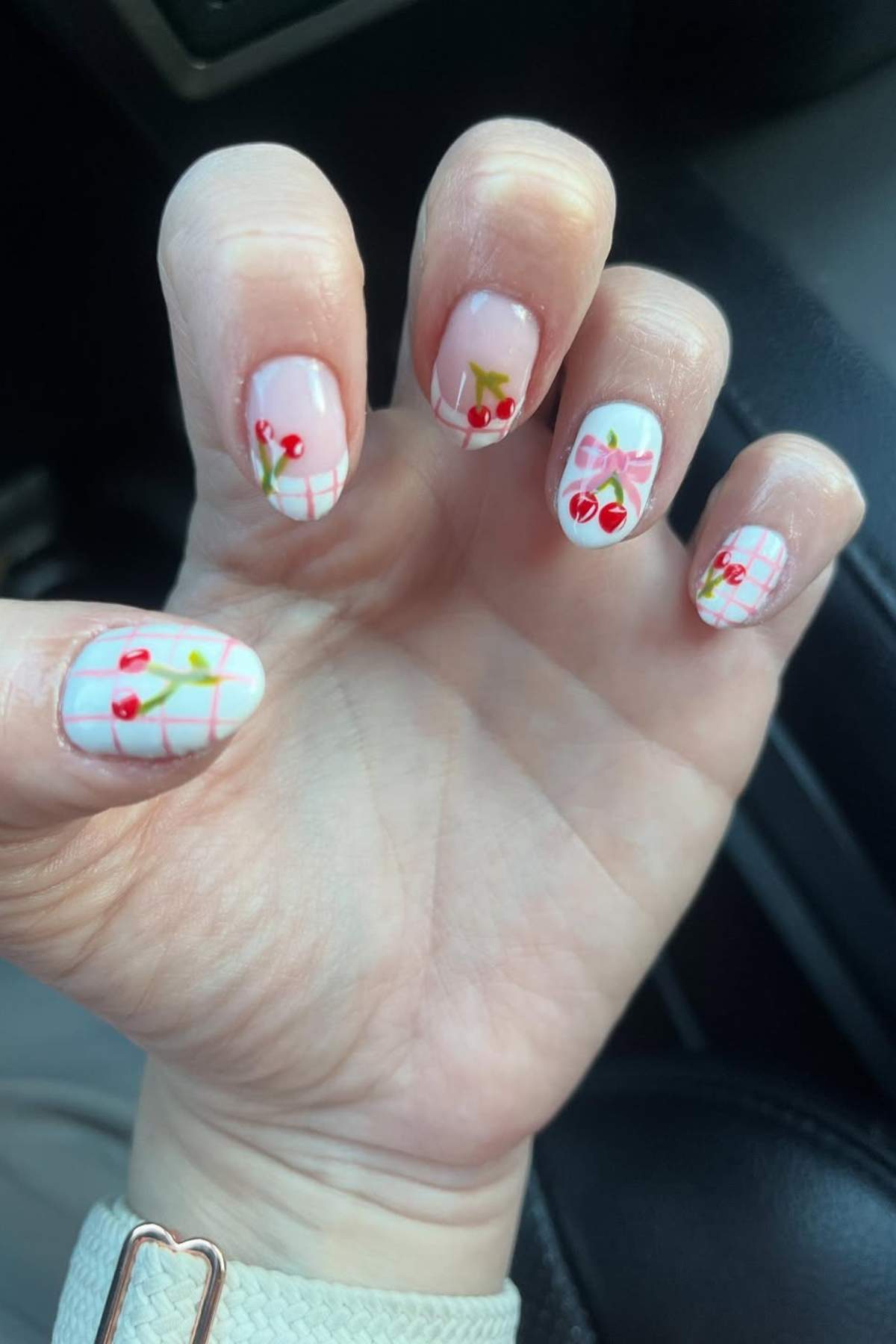 A hand with manicured nails featuring cherry designs and white grid patterns on a light background.