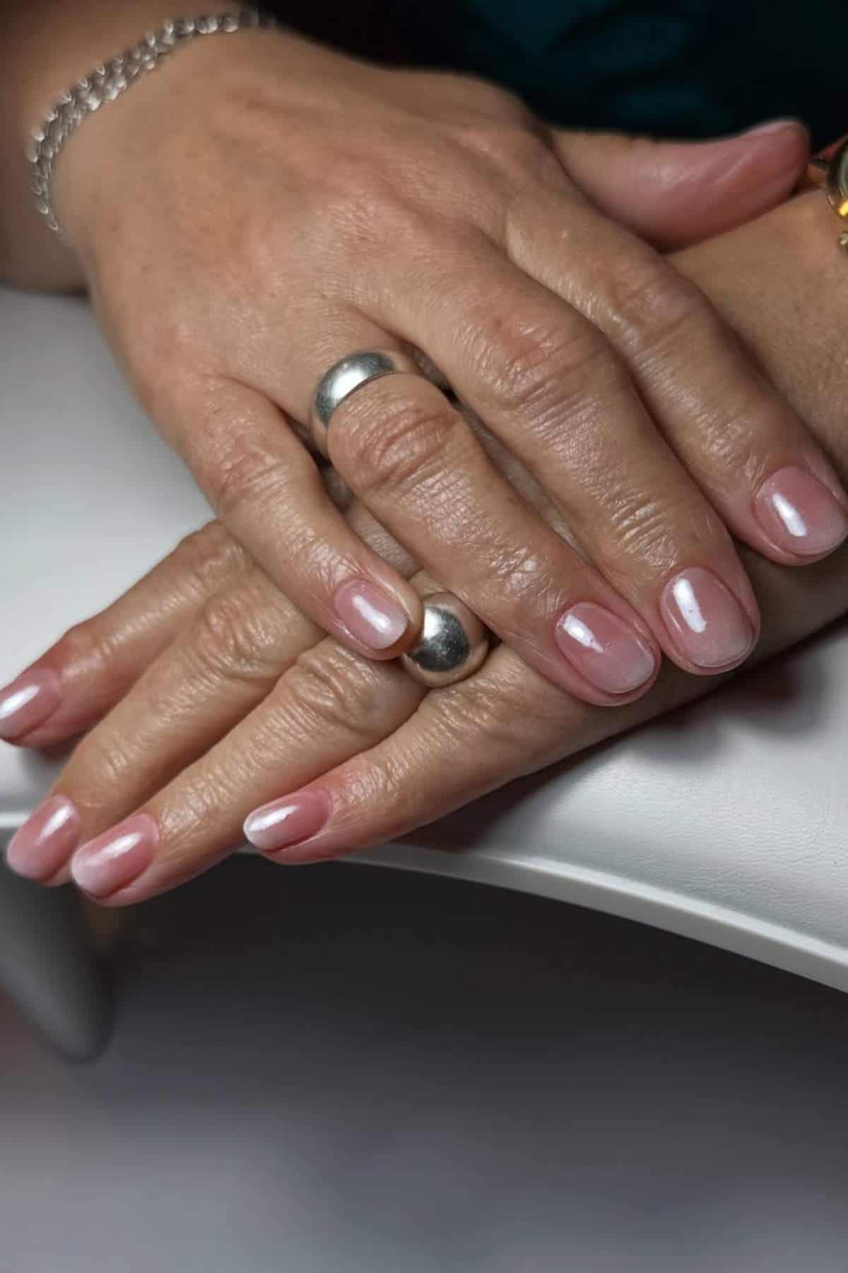 Close-up of two hands with short, oval-shaped nails polished in a light pink color. The hands are adorned with silver rings and a bracelet.