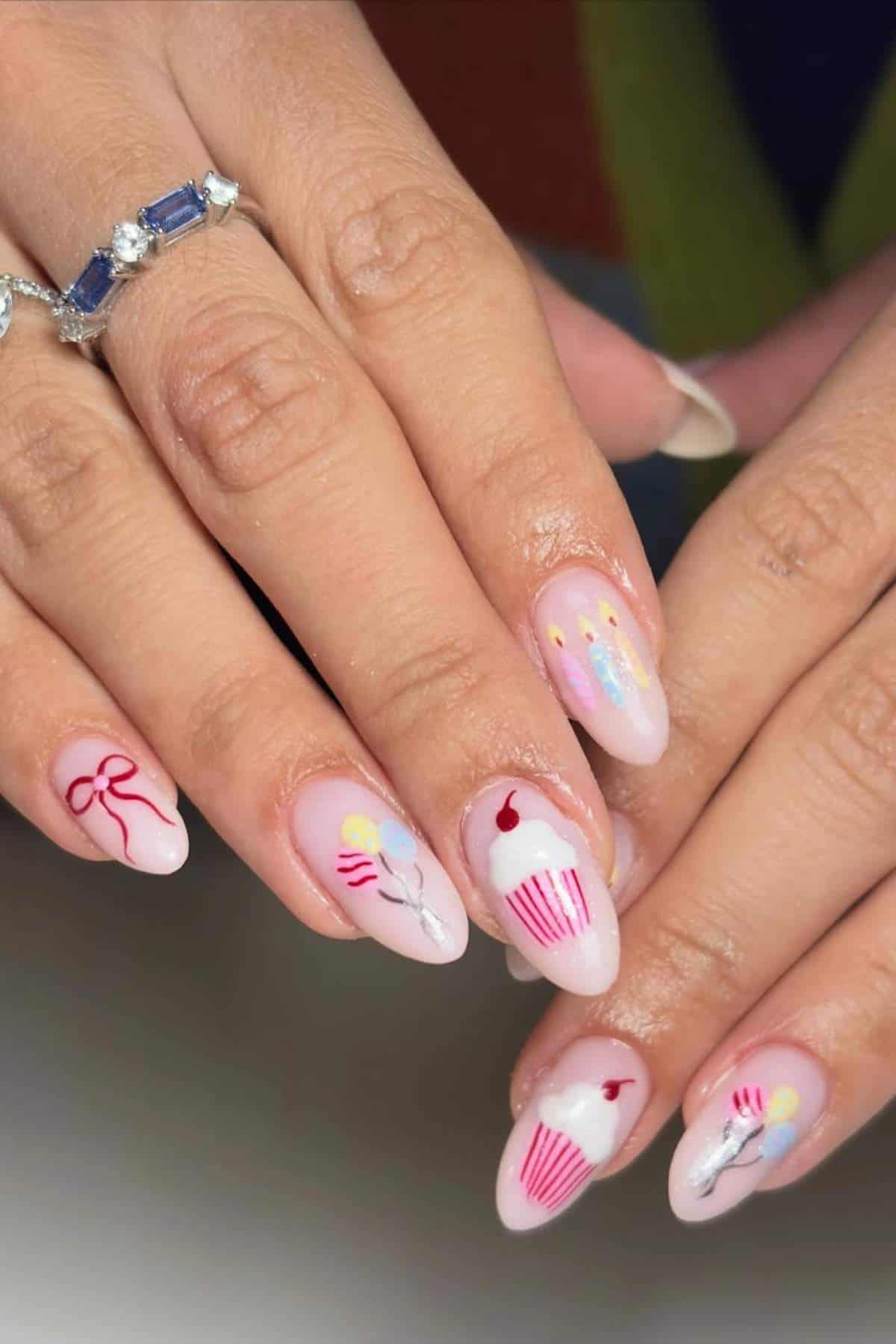 A hand with almond-shaped nails featuring birthday-themed nail art, including cupcakes, candles, balloons, and a bow. The person is wearing a blue gemstone ring.
