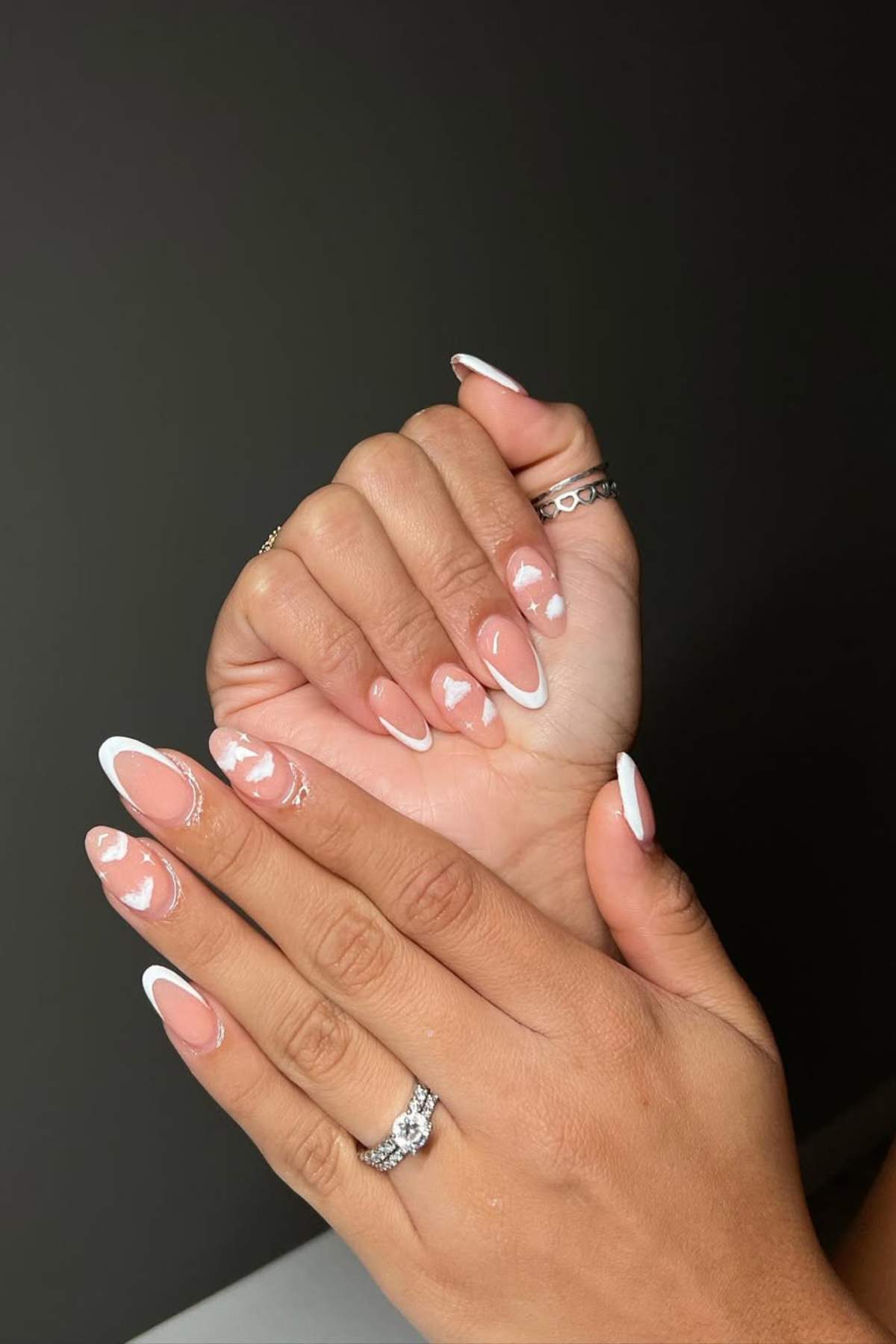 Hands with almond-shaped nails featuring a French manicure with white tips and small white heart designs on a neutral pink base. Multiple rings are worn on both hands.