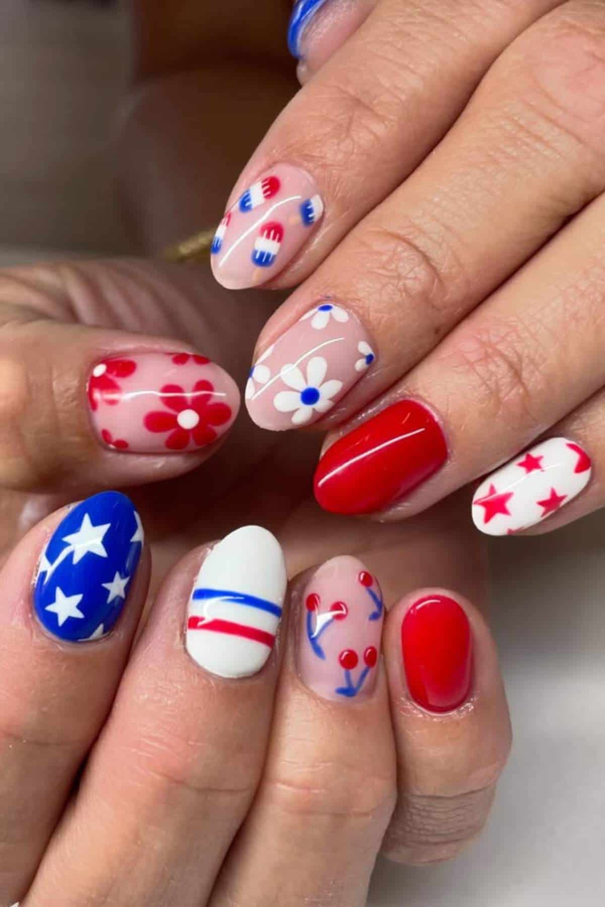 Hands with manicured nails featuring patriotic designs in red, white, and blue, including stars, stripes, flowers, and abstract patterns.