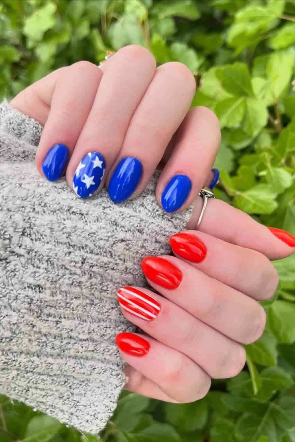 Hands with manicured nails; one hand has blue polish with white stars, the other has red polish with white stripes, resembling the American flag; green leaves in the background.