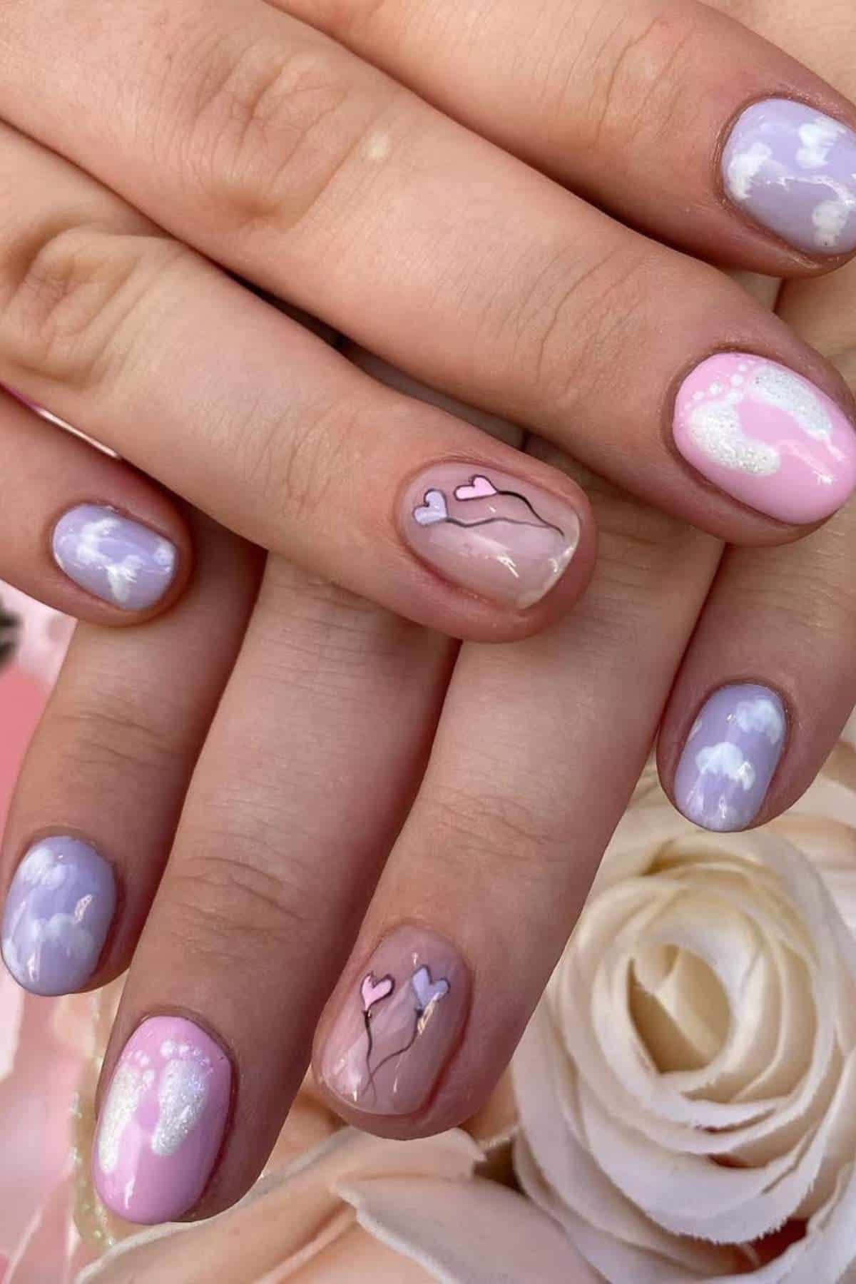 Close-up of hands with pastel-colored nail art featuring clouds, sparkles, and heart designs, set against a light floral background.