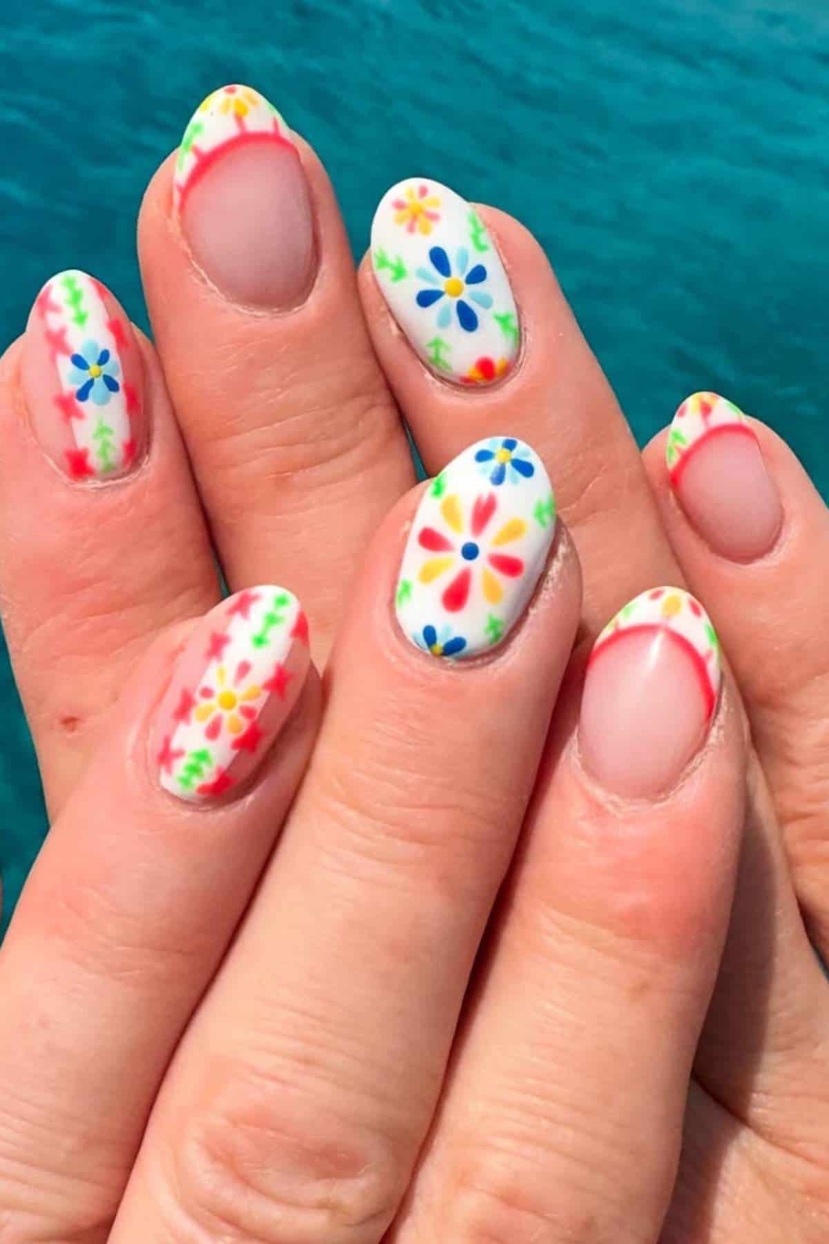 A hand with almond-shaped nails featuring a colorful floral pattern and French tips, with a blue background.