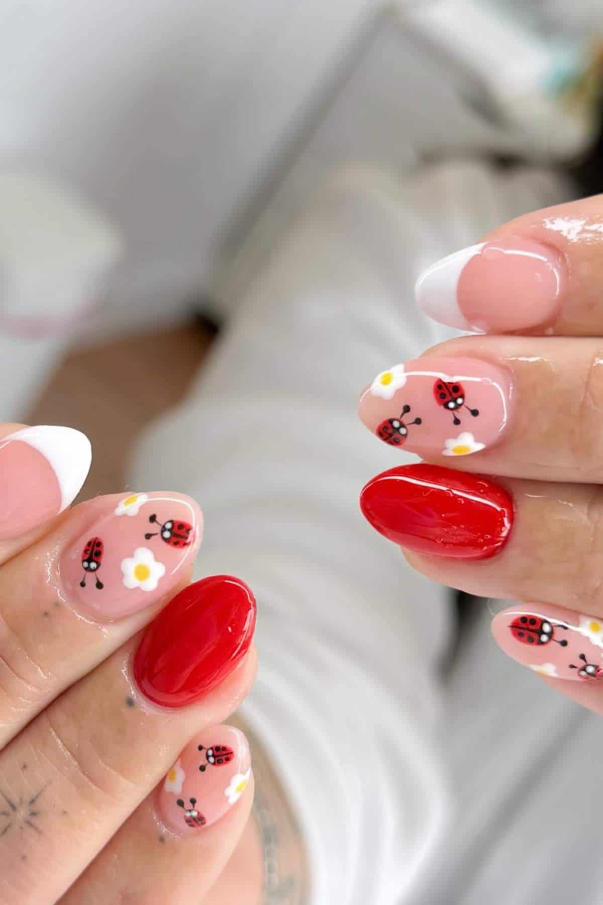 Close-up of manicured nails with red polish, French tips, and nail art featuring ladybugs and white daisies on a light pink base.