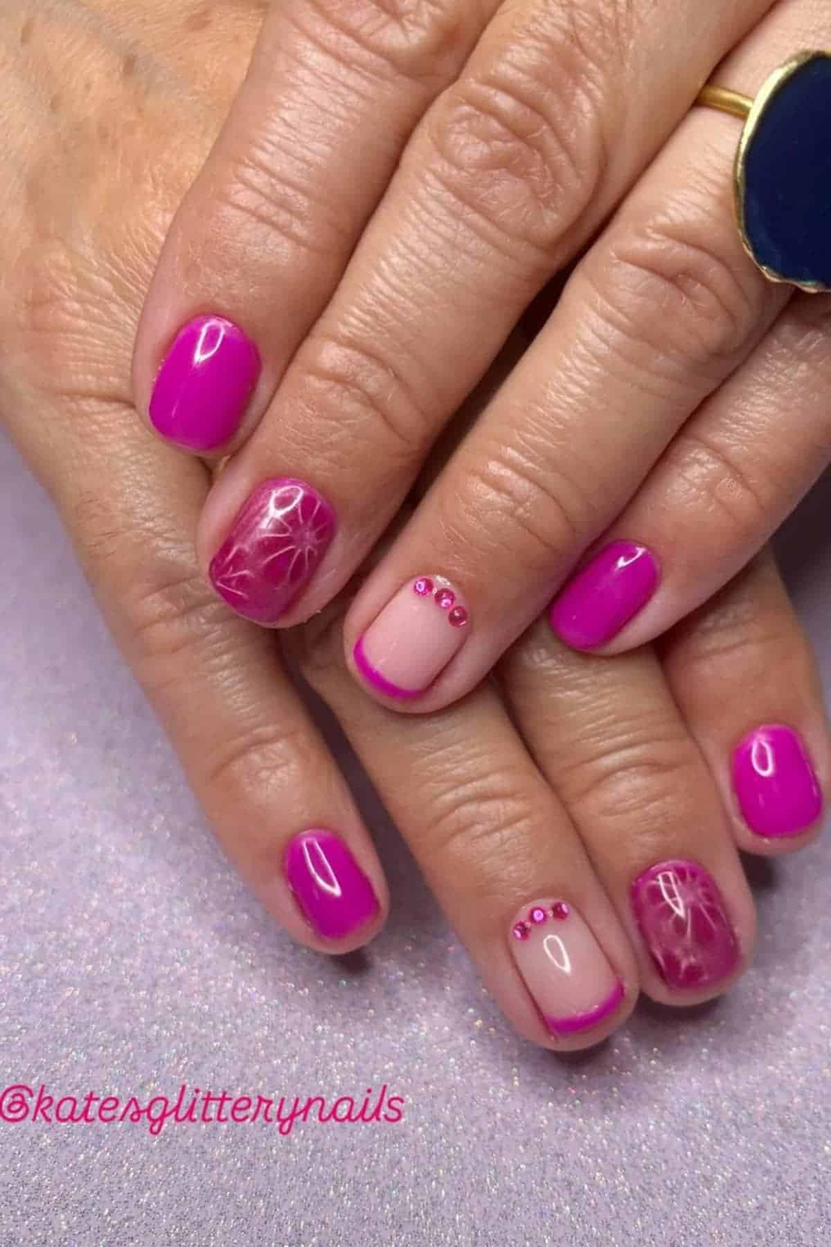 Hands with short, square nails painted pink; some nails have floral designs, pink tips, or pink rhinestone accents. The hand wears a gold ring with a large blue stone.