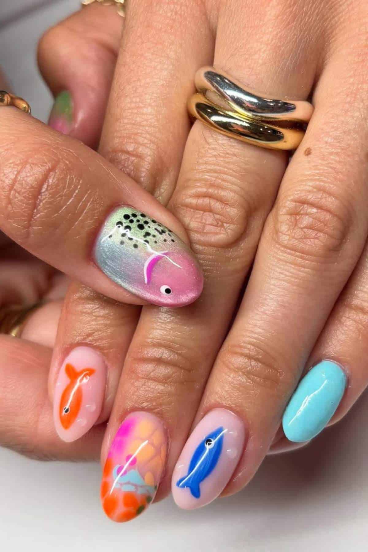 A hand with multicolored nail art featuring fish designs, scales, orange spots, and an accent nail painted light blue, with gold rings on the fingers.