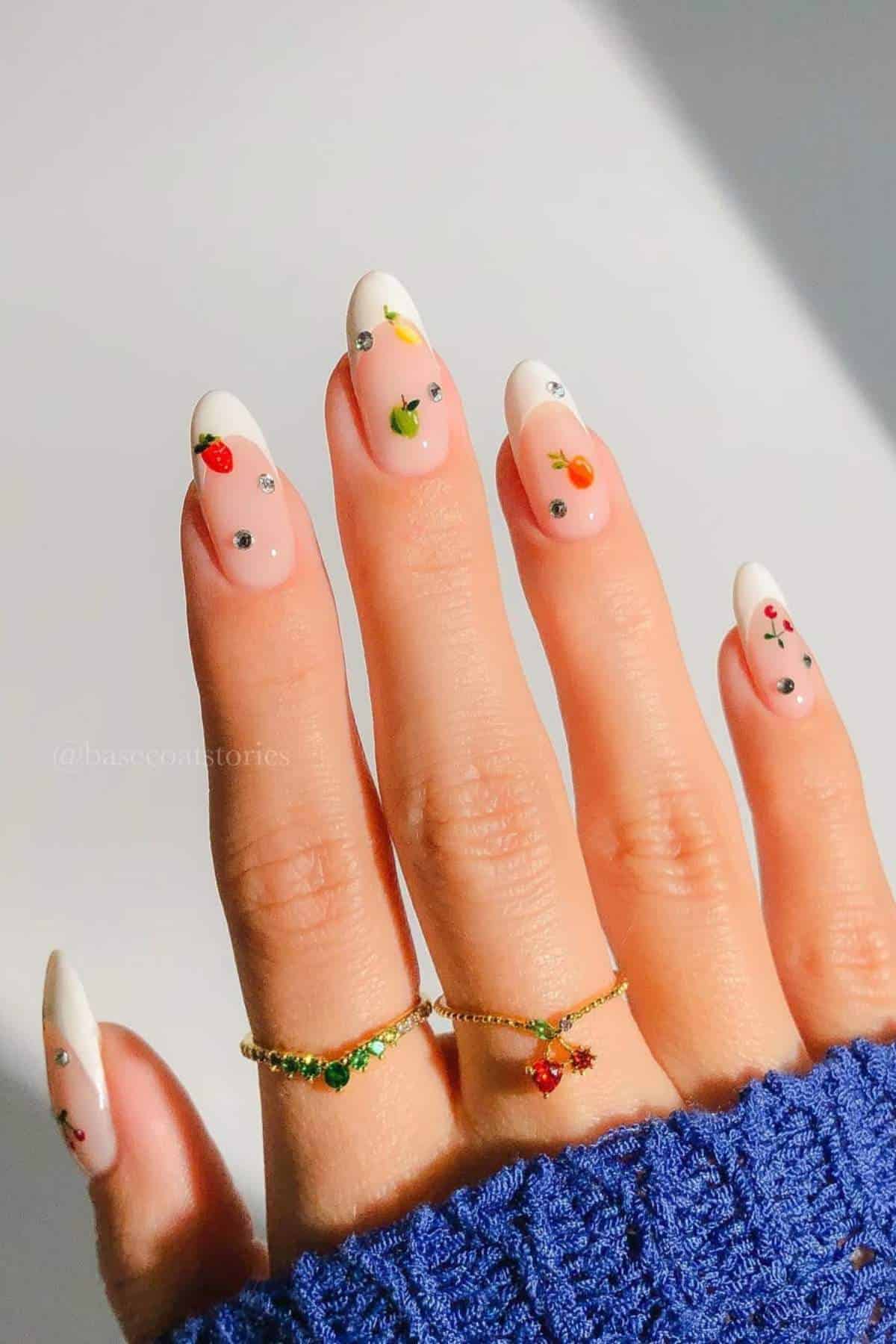 A hand with long, almond-shaped nails features a French manicure design with small fruit and gem decorations. The person wears two gold rings and a blue knit sleeve is visible.