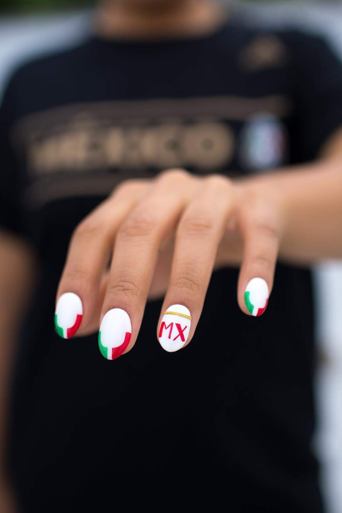 A hand with white-painted nails featuring red and green designs; the ring finger has "MX" and a yellow arch, referencing the Mexican flag and culture.