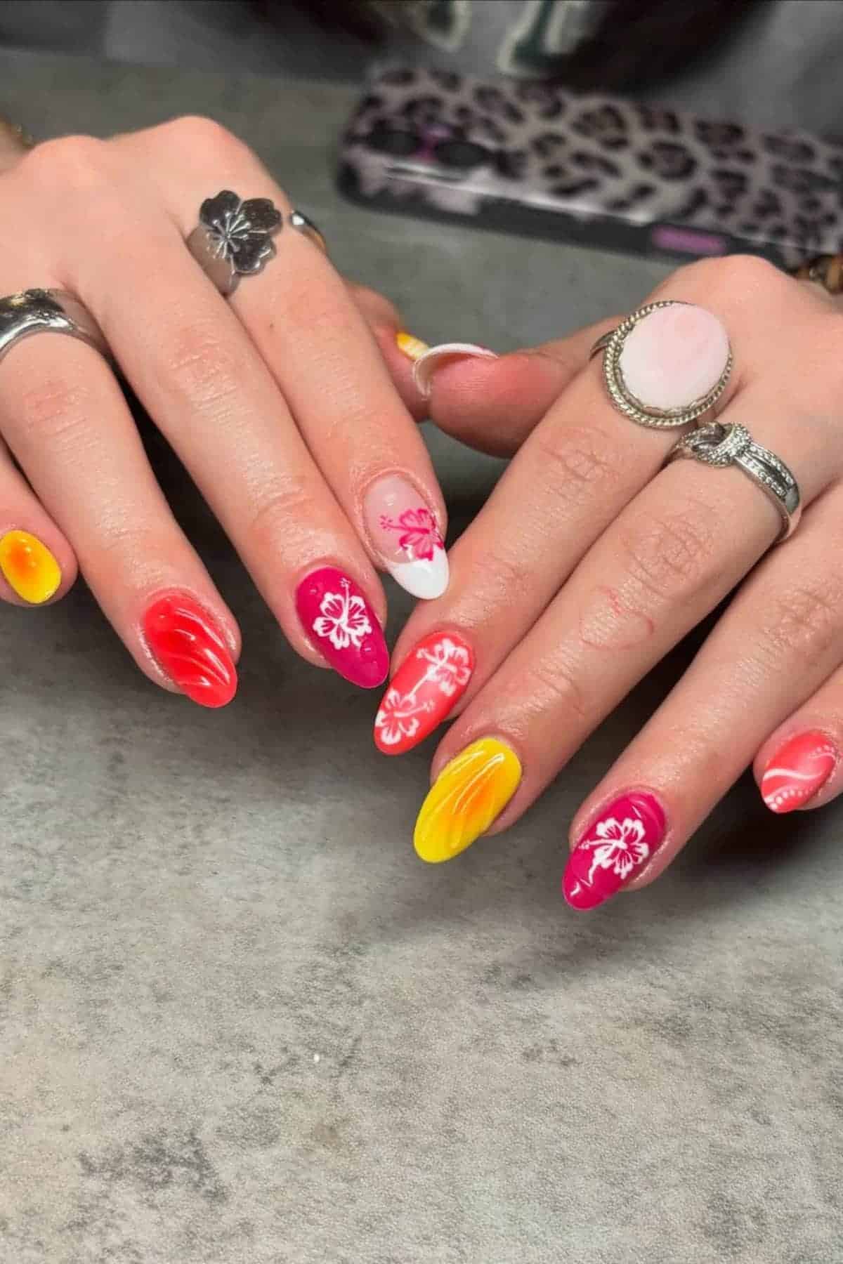 Hands with long, pointed nails painted in red, pink, orange, yellow, and white with floral designs. The person is wearing several rings.