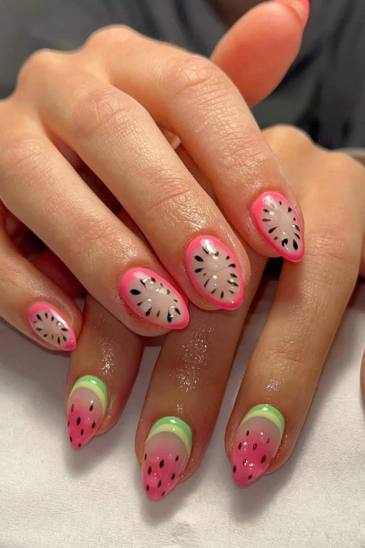 A close-up of hands with almond-shaped nails painted in a watermelon and kiwi fruit design with pink, green, black, and white colors.