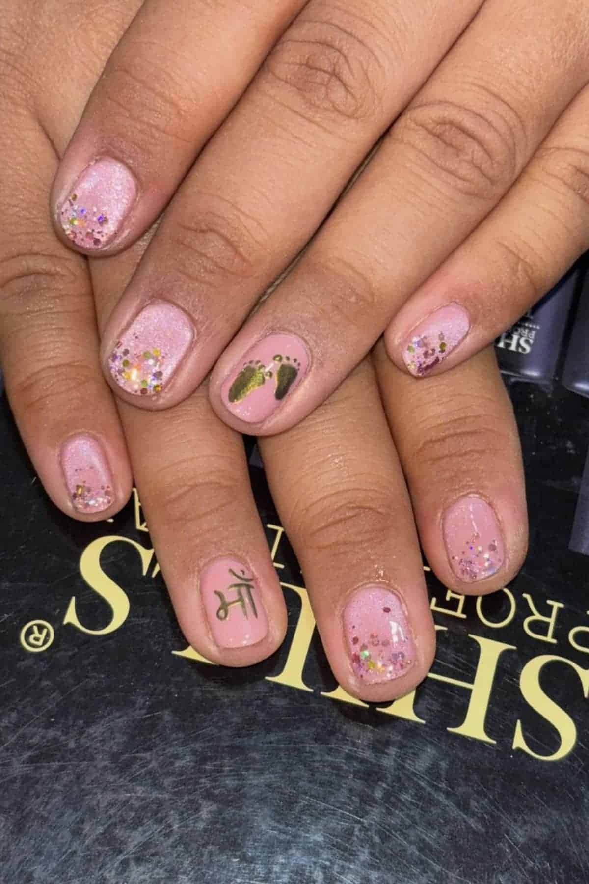Close-up of hands with short, light pink nails featuring glitter, small footprints, and a Hindi symbol as nail art, displayed over a black and yellow surface.
