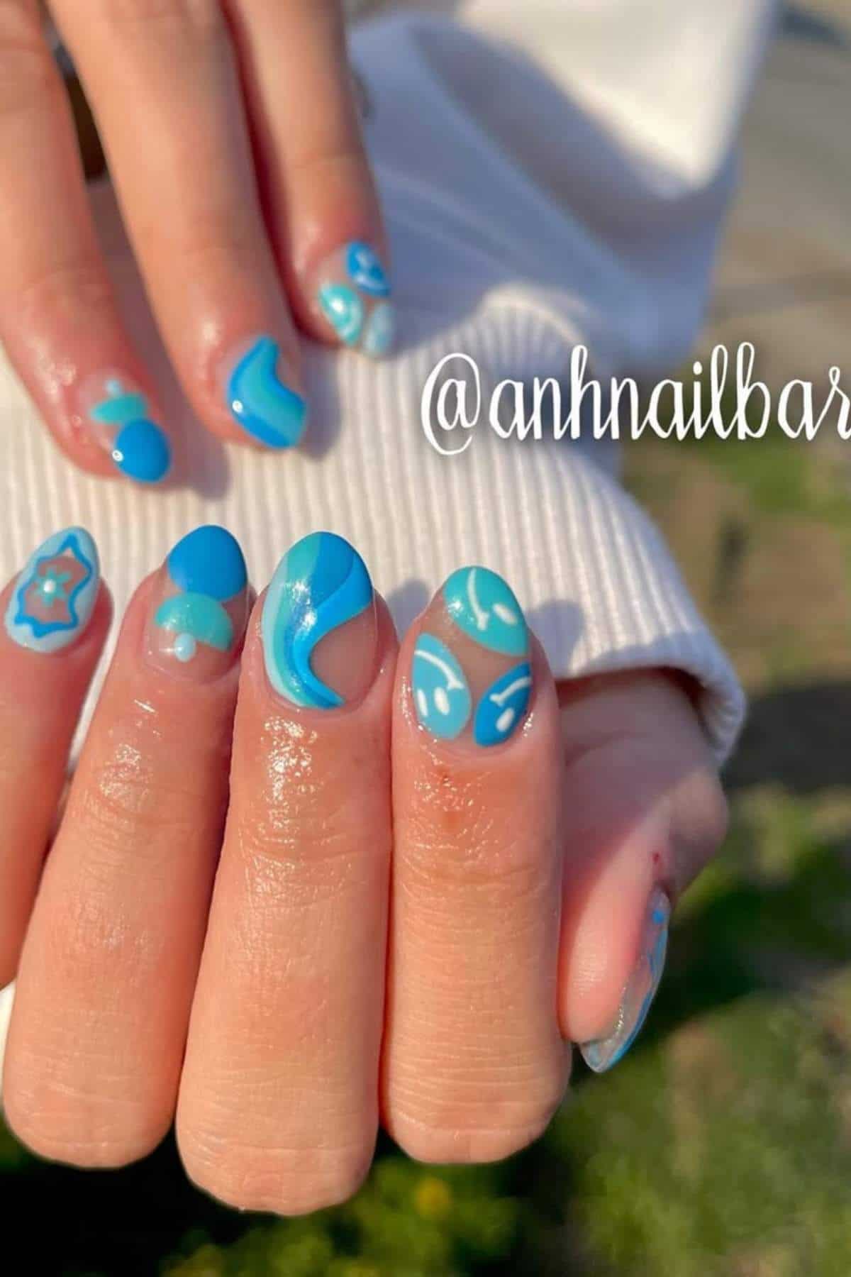 A hand with long almond-shaped nails features blue and turquoise abstract and floral designs on a glossy finish.