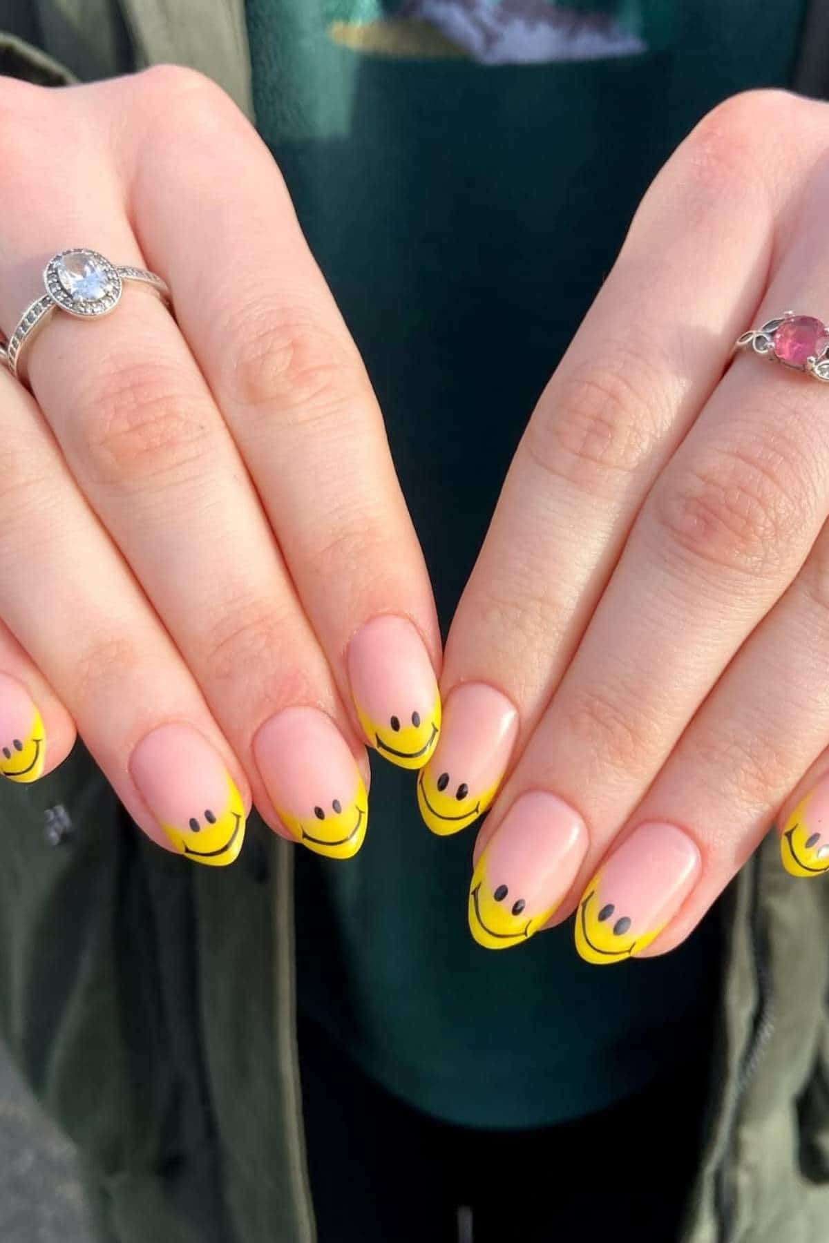 Close-up of hands with yellow French tip nails featuring black smiley face designs on each tip. The person is wearing two rings, one on each hand.