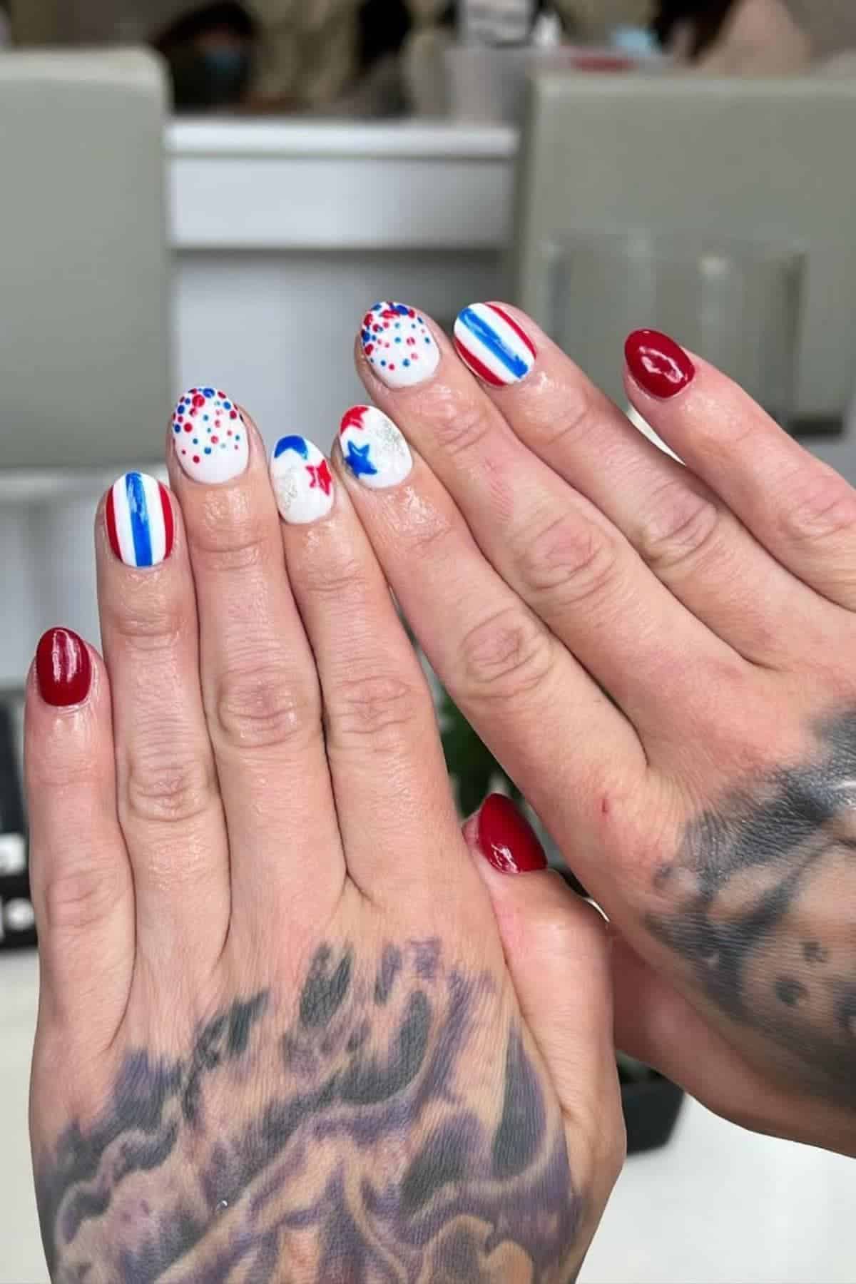 Hands with red, white, and blue nail art featuring dots, stripes, and stars. Some nails are solid red, others have patriotic designs. Visible tattoos on one hand.