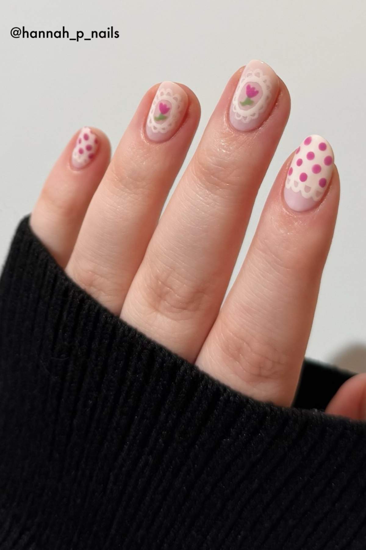 A hand with short nails featuring pink heart and polka dot nail art designs, partially covered by a black sleeve.