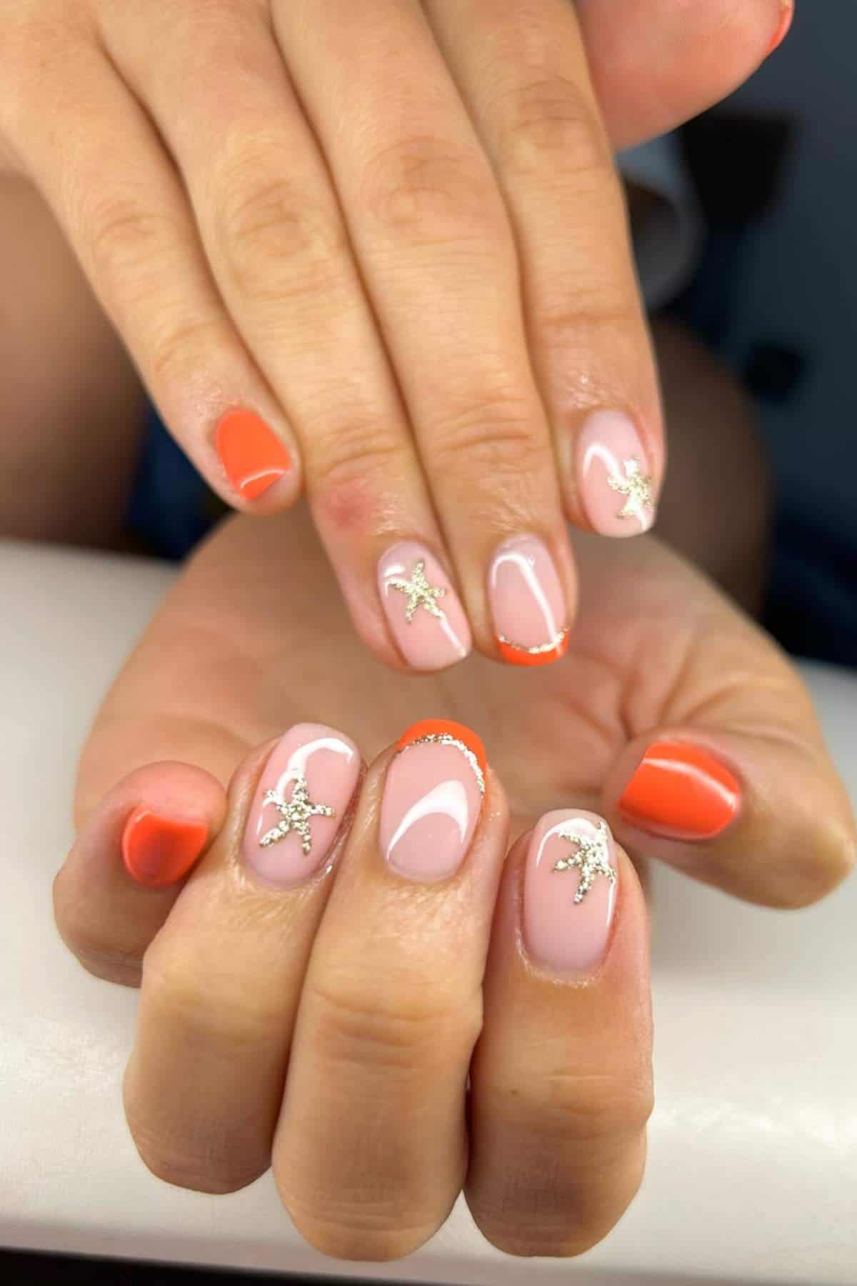 Hands with manicured nails featuring a mix of orange polish, nude base, and silver starfish charms as nail art accents.