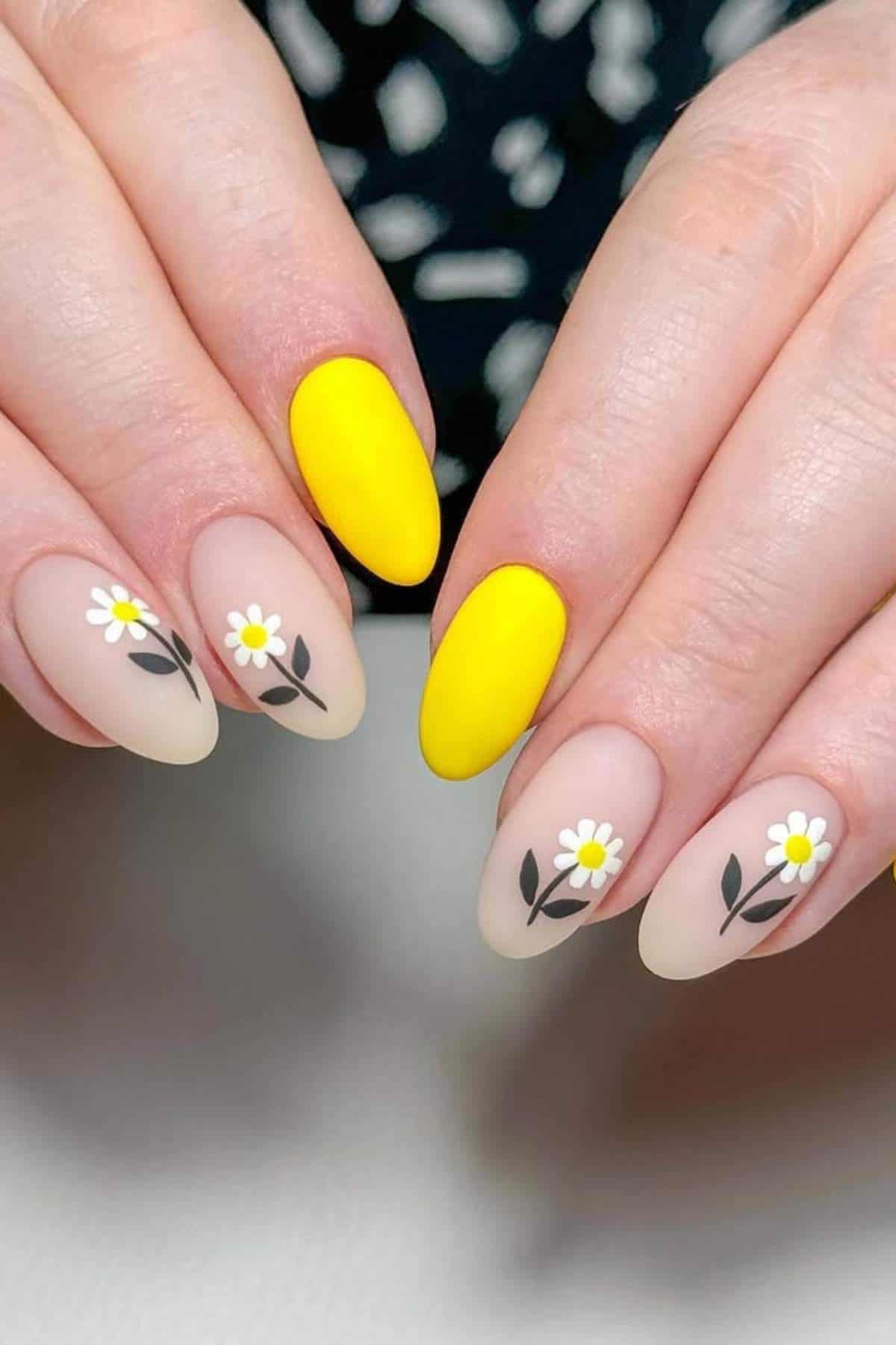 Hands with almond-shaped nails; two nails painted solid yellow and three with a nude base and daisy flower designs on each hand.